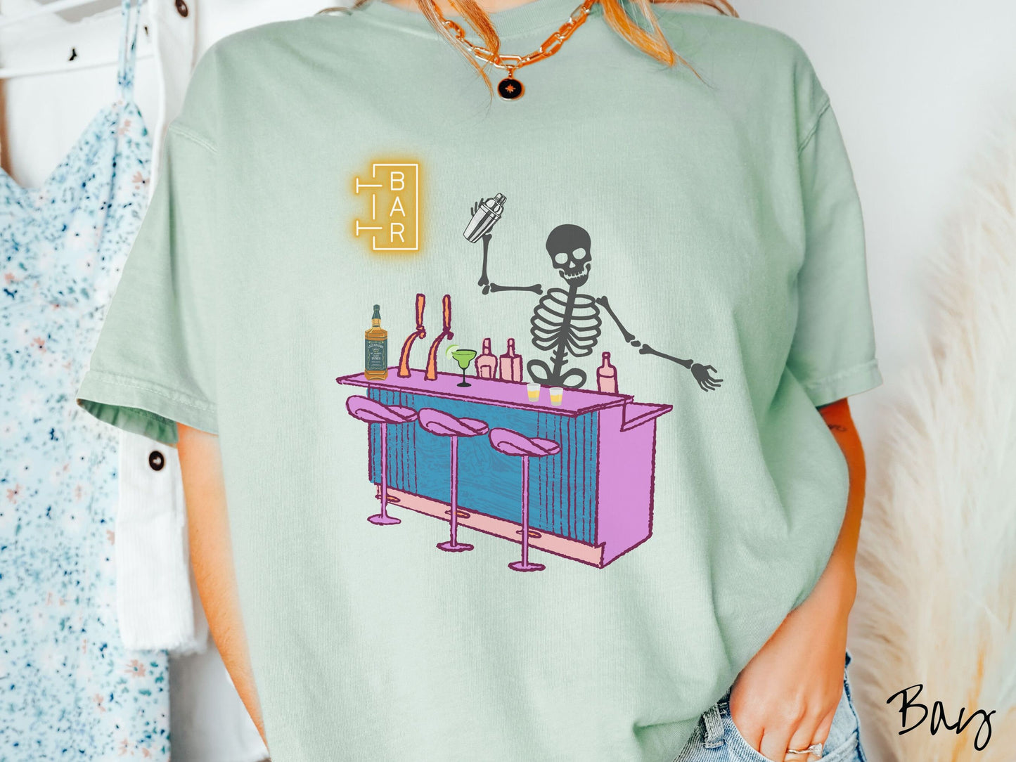A woman wearing a cute, vintage bay colored Comfort Colors t-shirt showing a skeleton bartender behind a purple bar mixing drinks. There is a yellow bar sign behind the bar and three purple chairs in front. There are liquor bottles on top.
