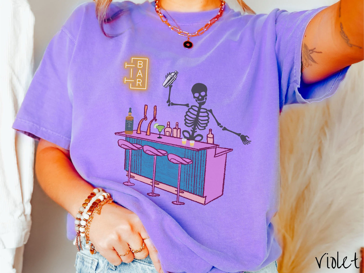 A woman wearing a cute, vintage violet colored Comfort Colors t-shirt showing a skeleton bartender behind a purple bar mixing drinks. There is a yellow bar sign behind the bar and three purple chairs in front. There are liquor bottles on top.