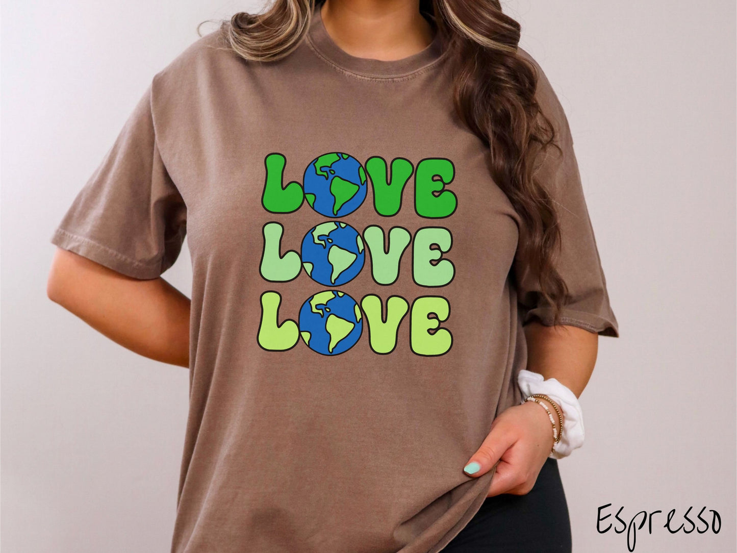 A woman wearing a cute, vintage espresso colored Comfort Colors shirt with the word Love listed vertically three times in varying shades of green, uppercase font. The Os in the words are pictures of the blue and green earth.