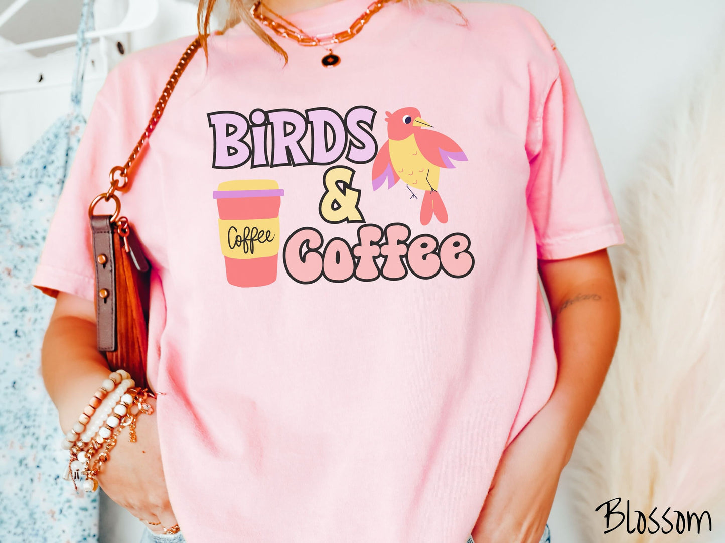 A woman wearing a cute, vintage blossom colored Comfort Colors shirt with the text Birds & Coffee in purple and light pink font. Next to the text is a red, yellow, and purple smiling bird in the air and a red and yellow to-go coffee cup.