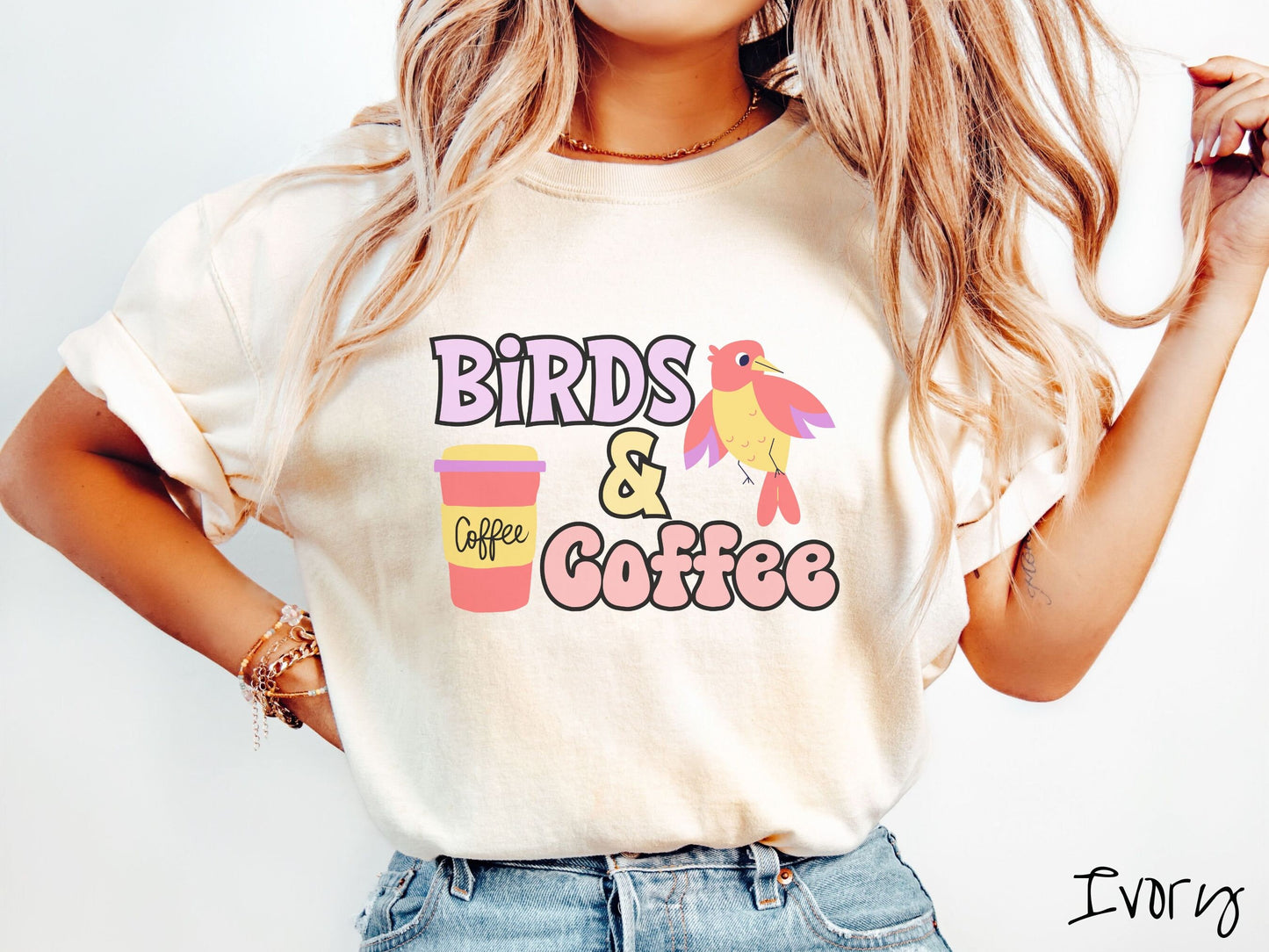 A woman wearing a cute, vintage ivory colored Comfort Colors shirt with the text Birds & Coffee in purple and light pink font. Next to the text is a red, yellow, and purple smiling bird in the air and a red and yellow to-go coffee cup.