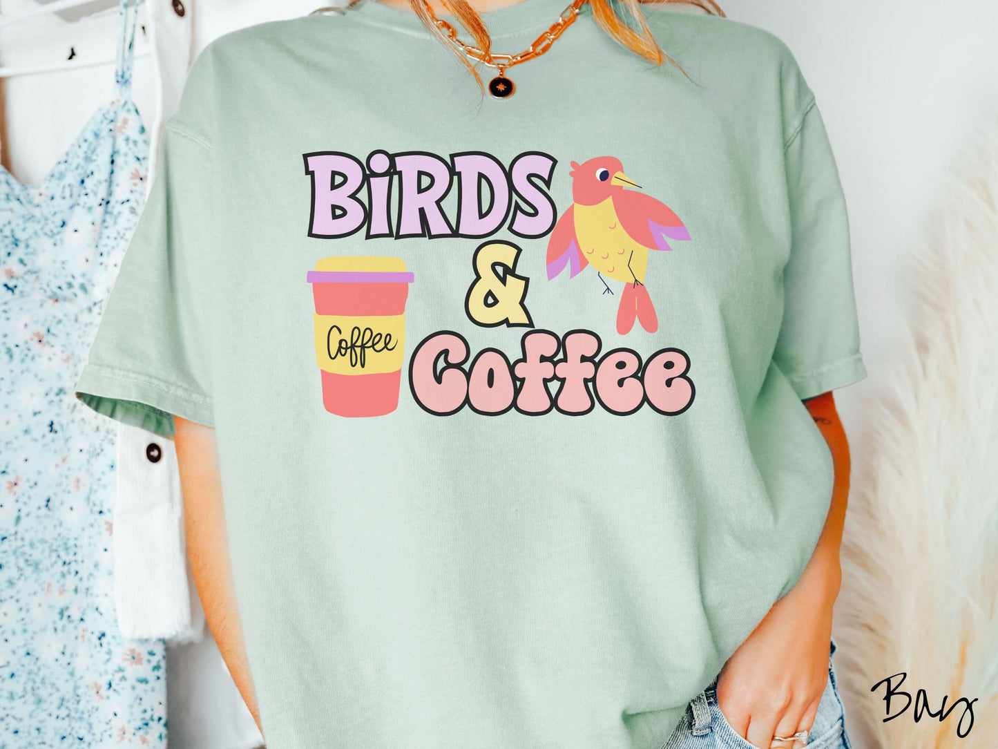 A woman wearing a cute, vintage bay colored Comfort Colors shirt with the text Birds & Coffee in purple and light pink font. Next to the text is a red, yellow, and purple smiling bird in the air and a red and yellow to-go coffee cup.