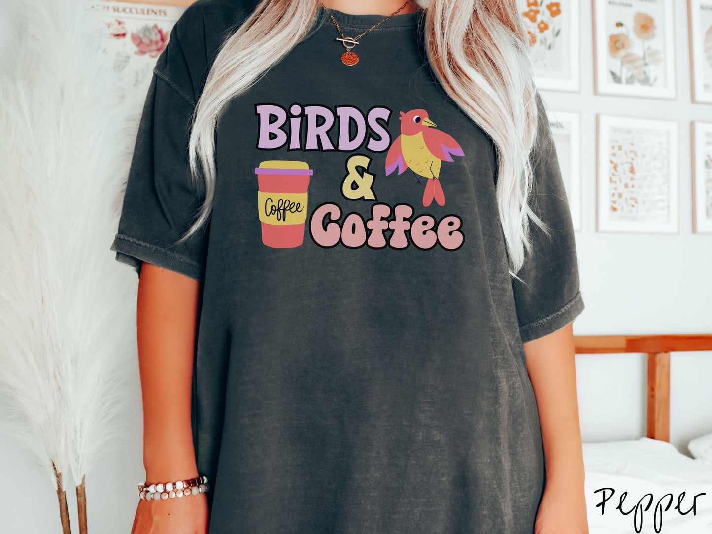A woman wearing a cute, vintage pepper colored Comfort Colors shirt with the text Birds & Coffee in purple and light pink font. Next to the text is a red, yellow, and purple smiling bird in the air and a red and yellow to-go coffee cup.