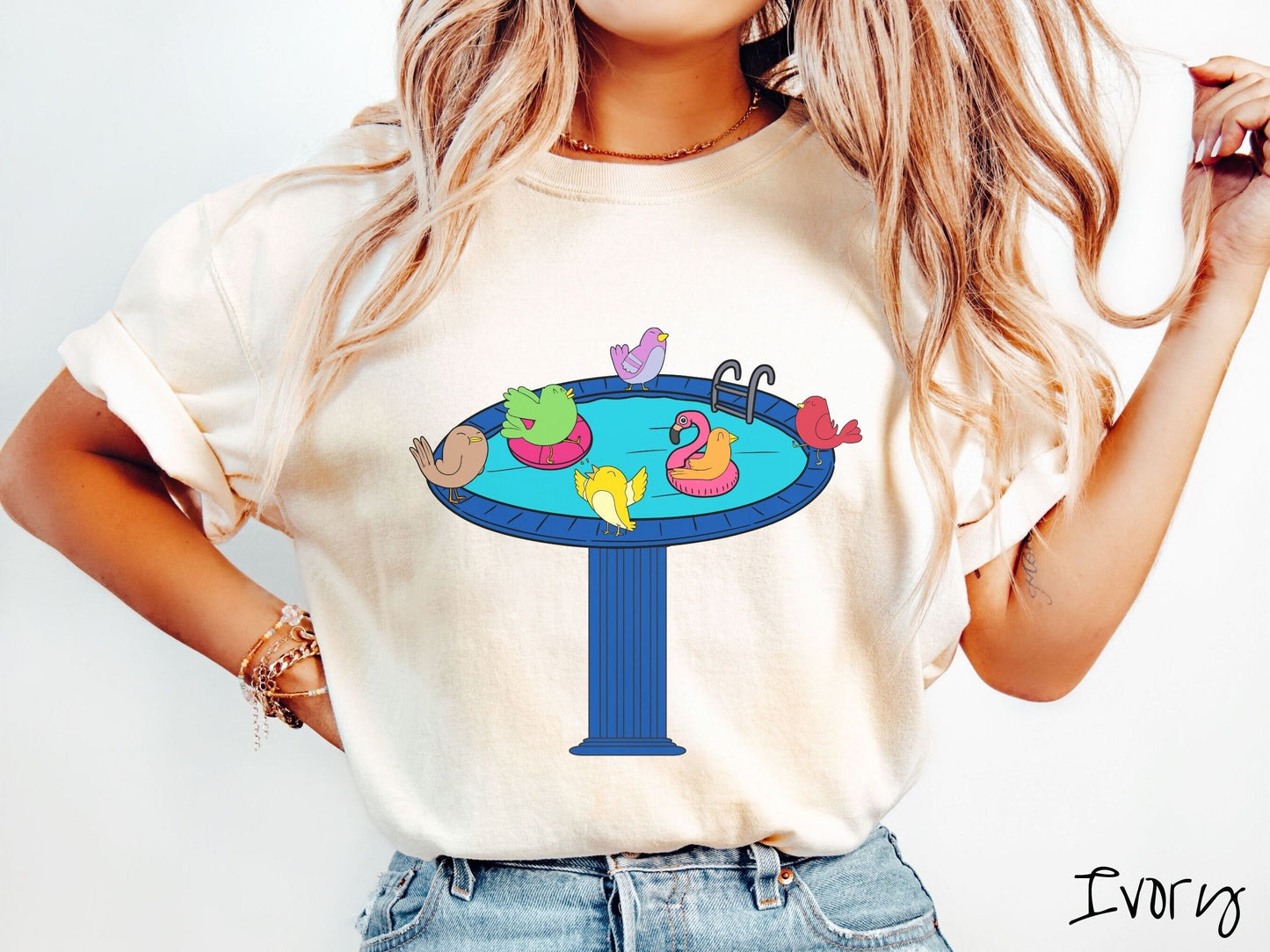 A woman wearing a cute, vintage pepper colored Comfort Colors shirt with a blue birdbath with a pool ladder filled with crystal clear blue water. In the water are colorful birds floating on colorful pool inflatables and enjoying the sun.