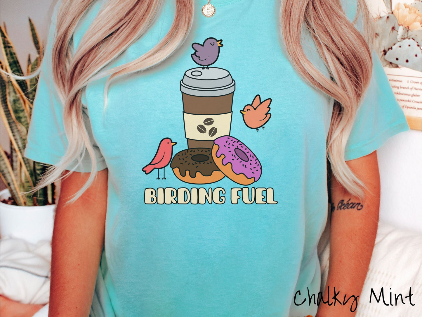 A woman wearing a cute, vintage chalky mint colored Comfort Colors shirt with a brown coffee cup next to a chocolate frosted donut and pink frosted donut. There are three orange, red, and purple birds sitting on the coffee cup and flying around.