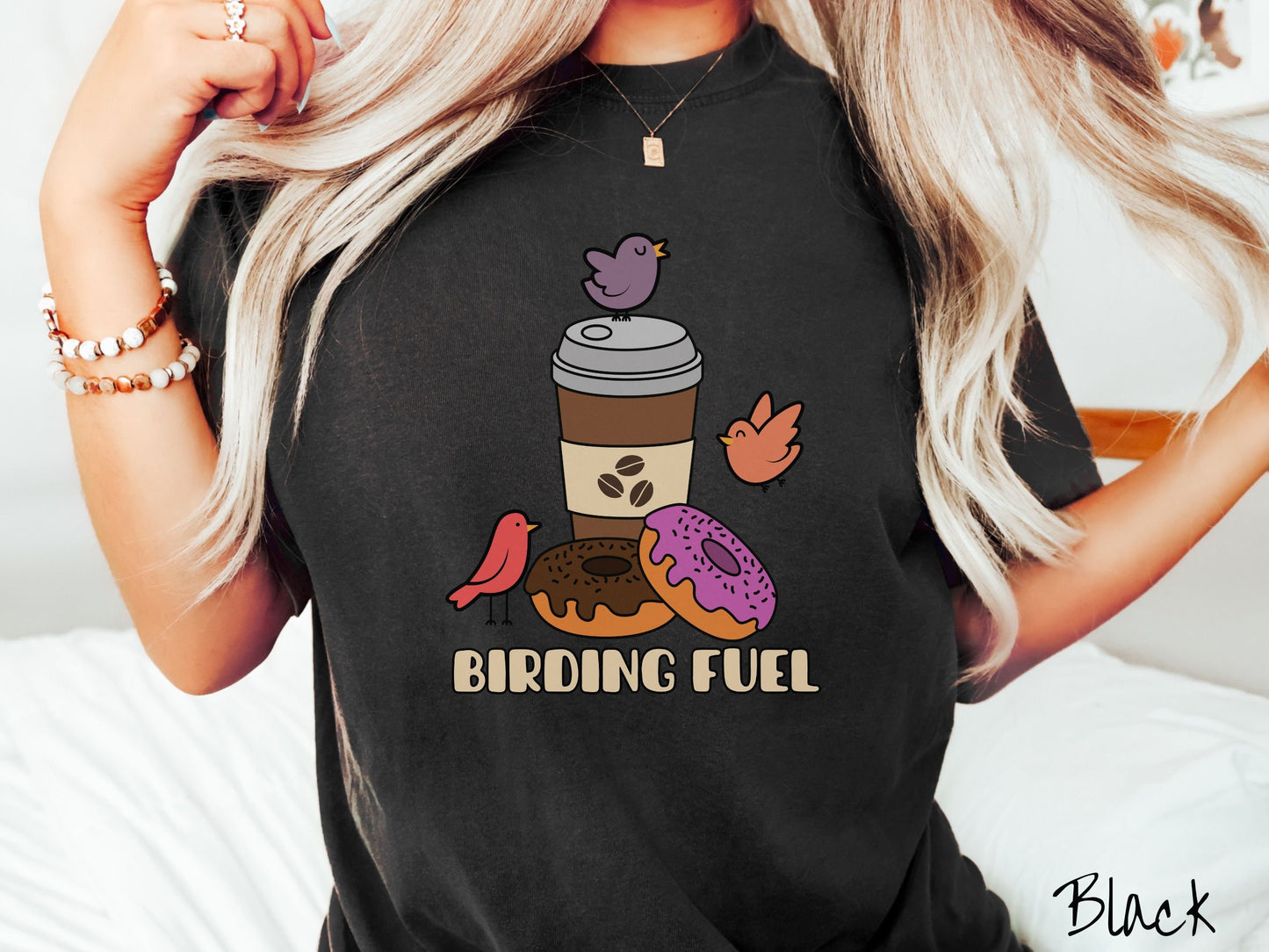 A woman wearing a cute, vintage black colored Comfort Colors shirt with a brown to-go coffee cup next to a chocolate frosted donut and pink frosted donut. There are three orange, red, and purple birds sitting on the coffee cup and flying around.