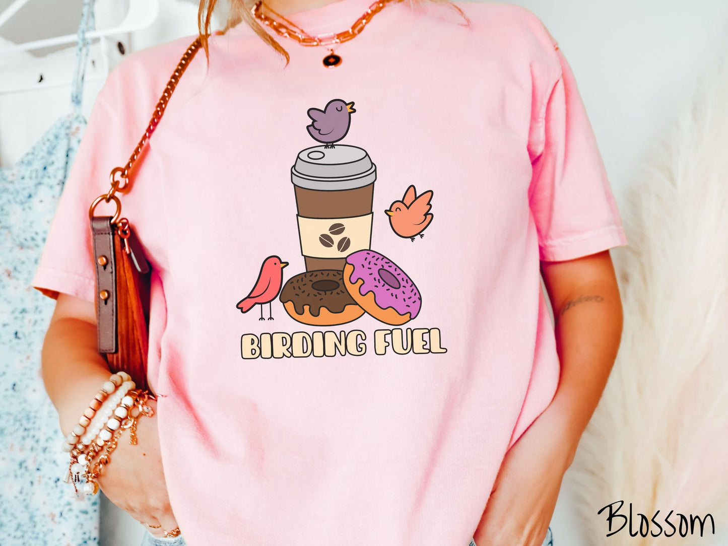 A woman wearing a cute, vintage blossom colored Comfort Colors shirt with a brown to-go coffee cup next to a chocolate frosted donut and pink frosted donut. There are three orange, red, and purple birds sitting on the coffee cup and flying around.