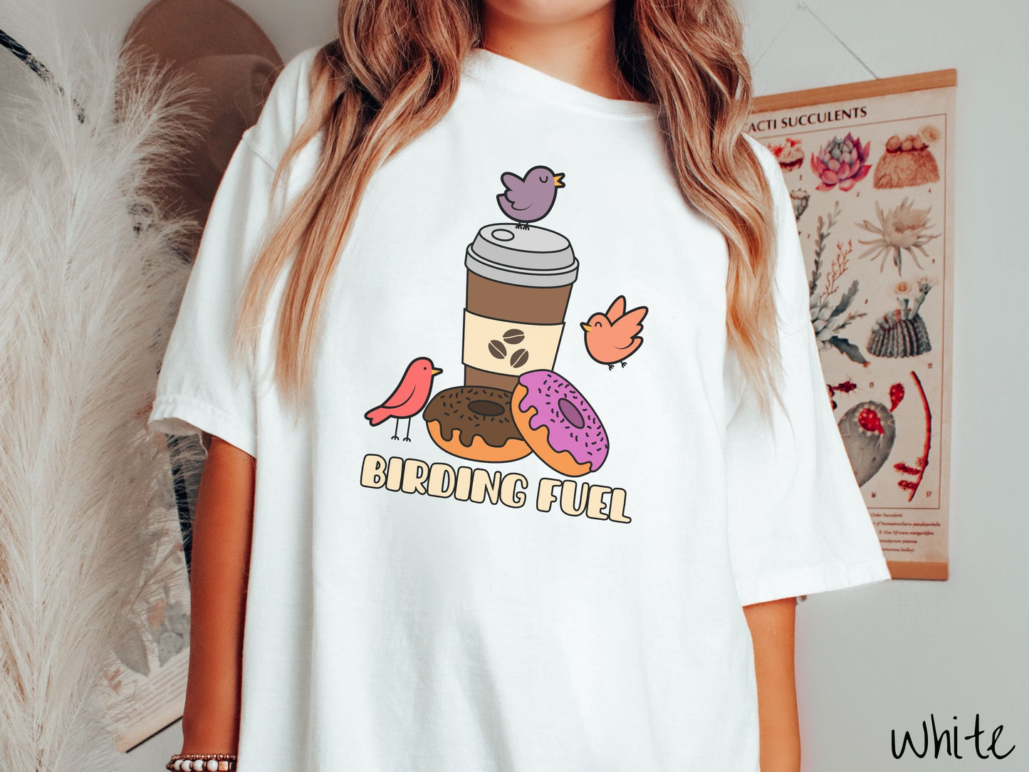 A woman wearing a cute, vintage white colored Comfort Colors shirt with a brown to-go coffee cup next to a chocolate frosted donut and pink frosted donut. There are three orange, red, and purple birds sitting on the coffee cup and flying around.