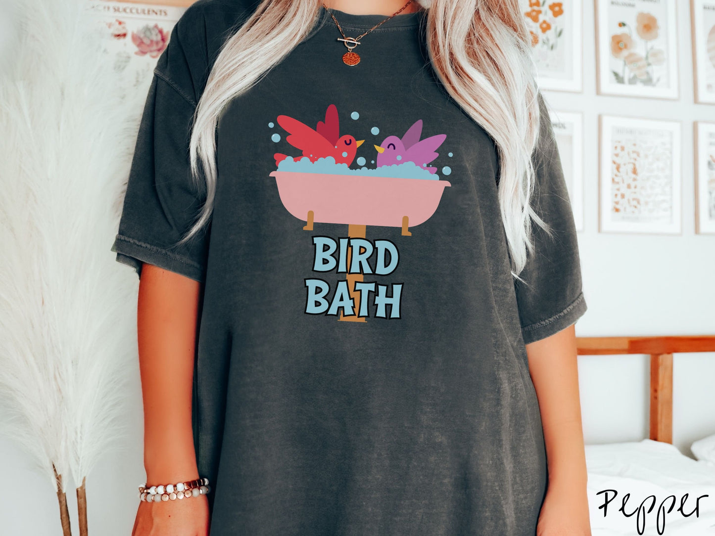 A woman wearing a cute, vintage pepper colored Comfort Colors shirt with a pink bathtub with a red bird and purple bird enjoying a bubble bath together. Below the bathtub are the words Bird Bath in light blue, uppercase font.