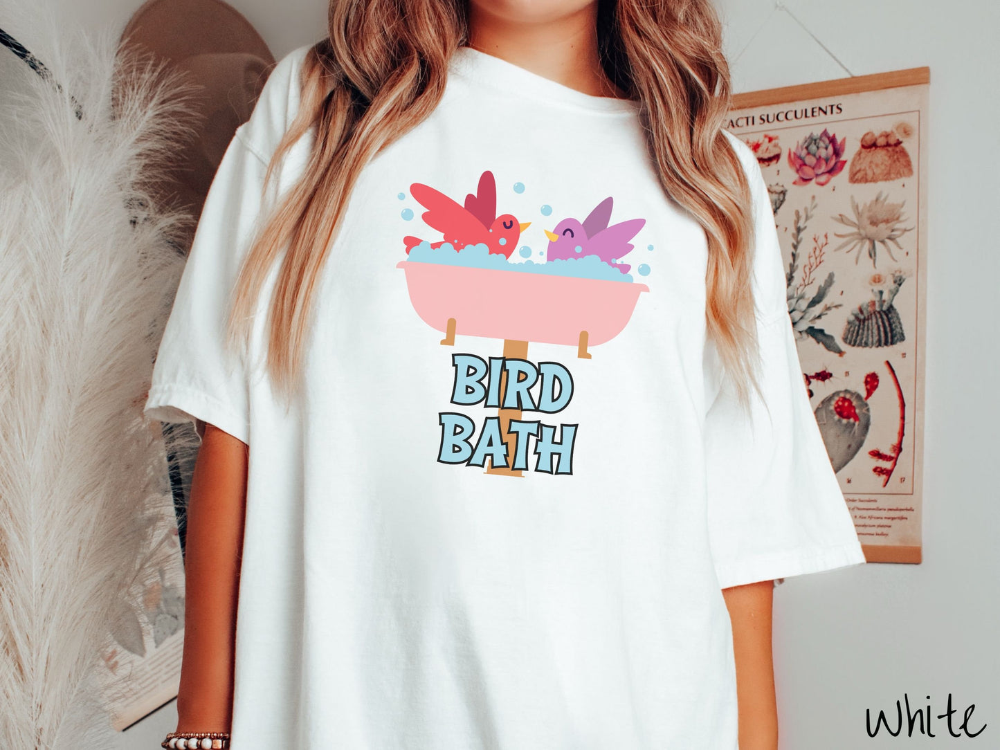 A woman wearing a cute, vintage white colored Comfort Colors shirt with a pink bathtub with a red bird and purple bird enjoying a bubble bath together. Below the bathtub are the words Bird Bath in light blue, uppercase font.