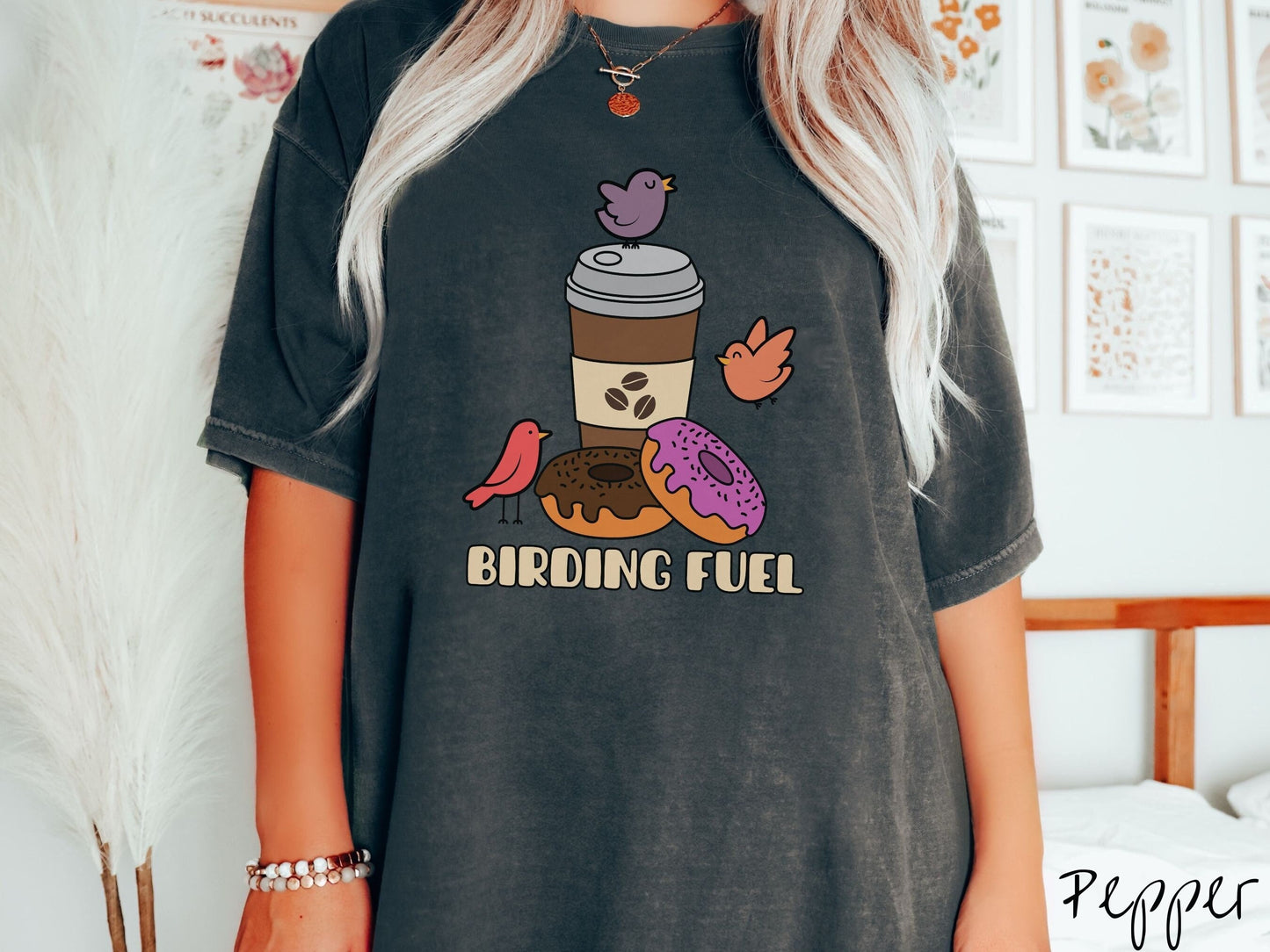 A woman wearing a cute, vintage pepper colored Comfort Colors shirt with a brown to-go coffee cup next to a chocolate frosted donut and pink frosted donut. There are three orange, red, and purple birds sitting on the coffee cup and flying around.
