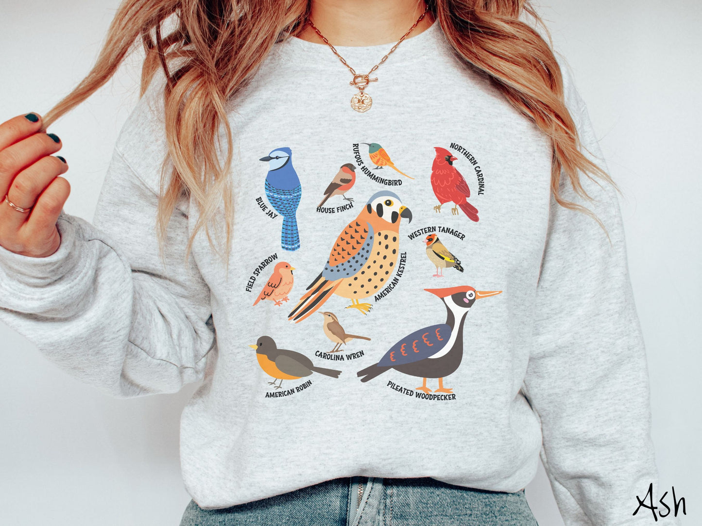 A woman wearing a cute, vintage ash colored comfy sweatshirt with pictures of nine different bird species and their names listed across the front. Some examples are the Blue Jay, House Finch, Western Tanager, and Carolina Wren.