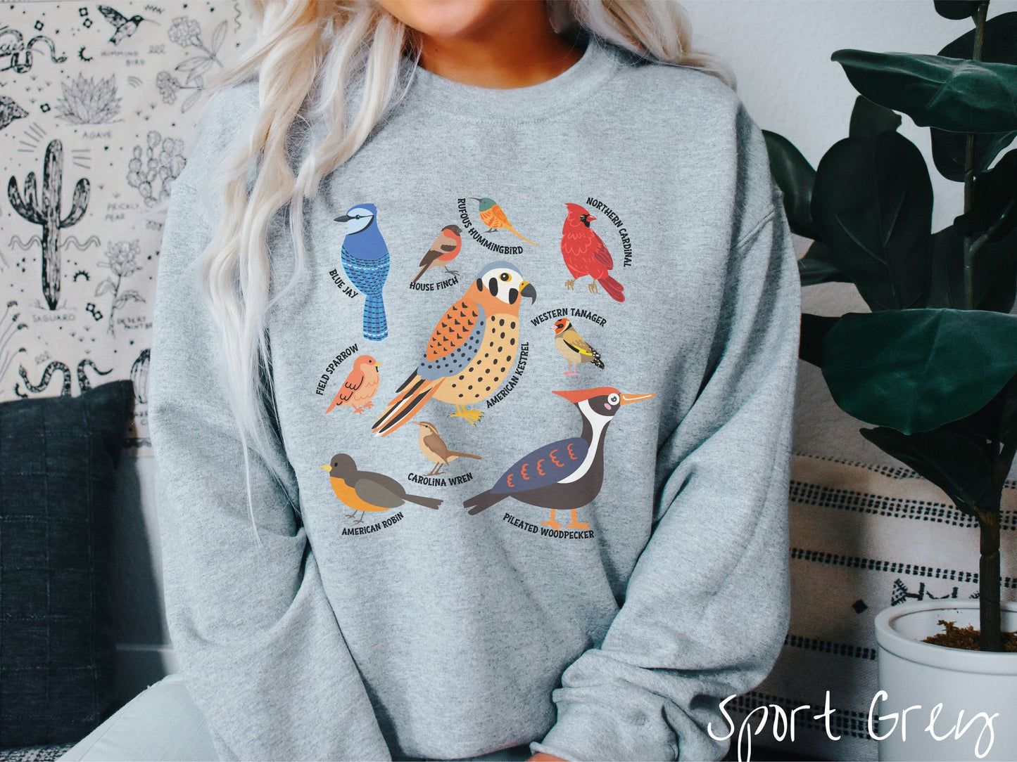 A woman wearing a cute, vintage sport grey colored comfy sweatshirt with pictures of nine different bird species and their names listed across the front. Some examples are the Blue Jay, House Finch, Western Tanager, and Carolina Wren.