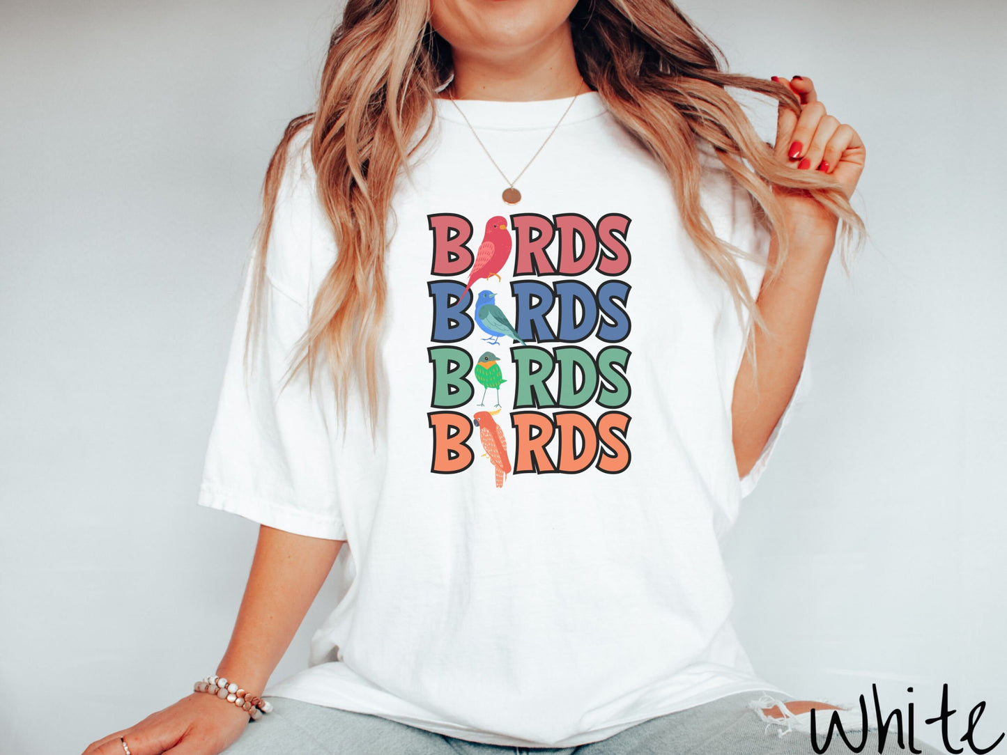 A woman wearing a cute, vintage white colored Comfort Colors t-shirt with the word BIRDS listed four times vertically in red, blue, green, and orange uppercase font. The Is in BIRDS are different kinds of colorful birds.