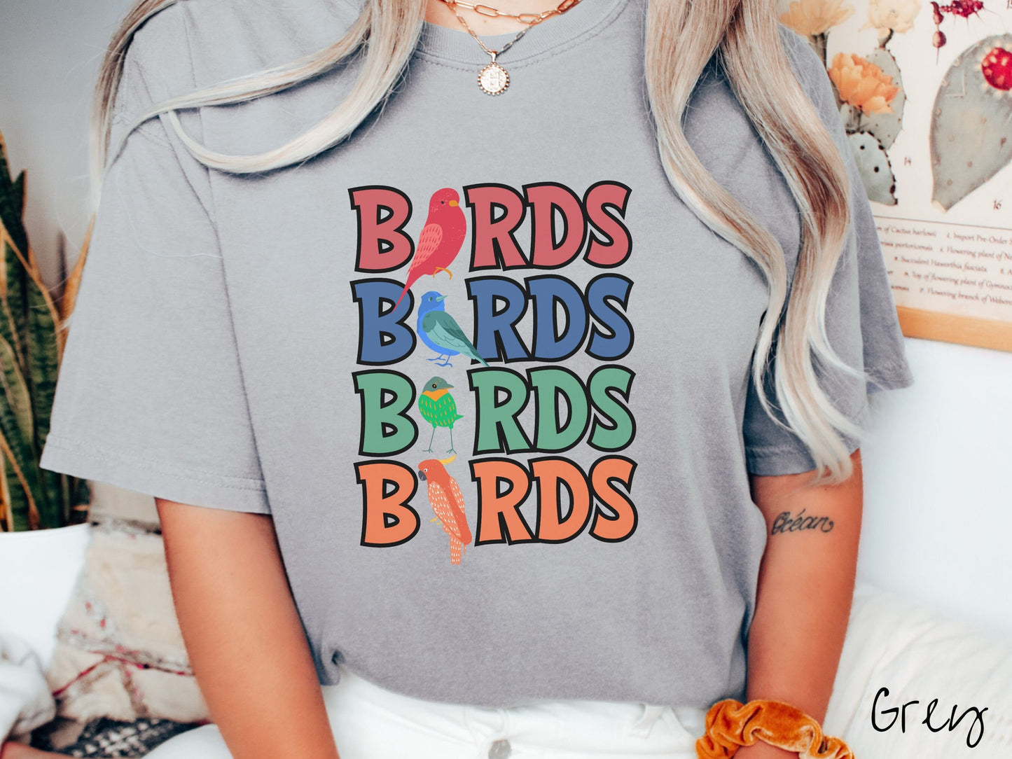 A woman wearing a cute, vintage grey colored Comfort Colors t-shirt with the word BIRDS listed four times vertically in red, blue, green, and orange uppercase font. The Is in BIRDS are different kinds of colorful birds.