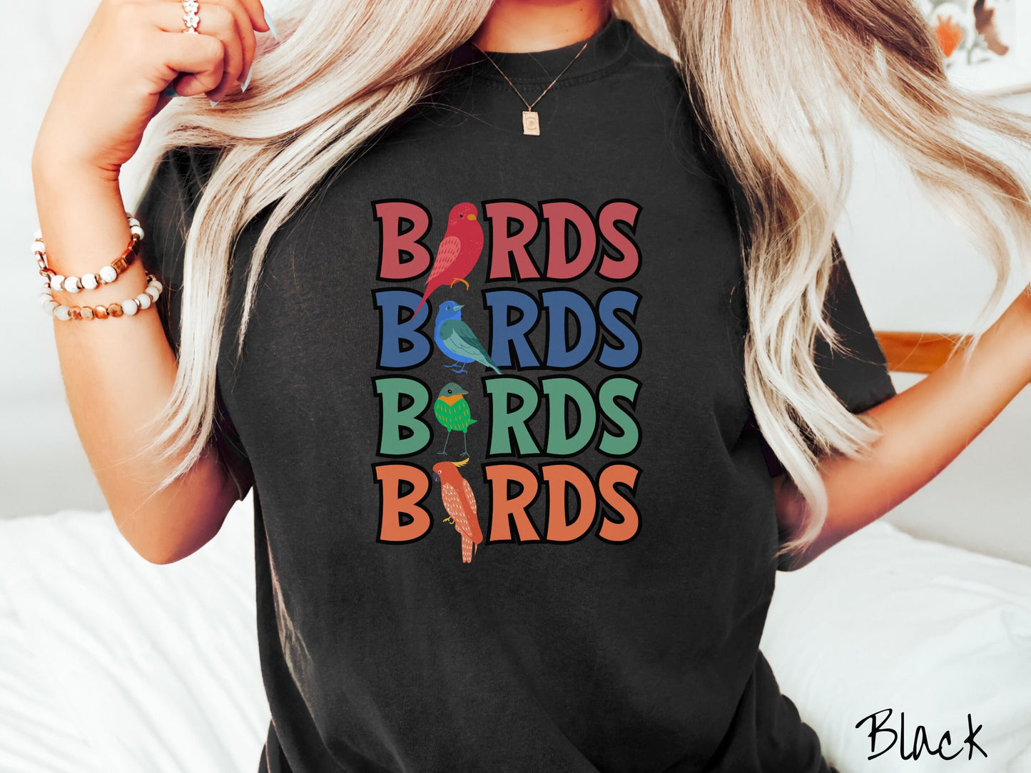A woman wearing a cute, vintage black colored Comfort Colors t-shirt with the word BIRDS listed four times vertically in red, blue, green, and orange uppercase font. The Is in BIRDS are different kinds of colorful birds.