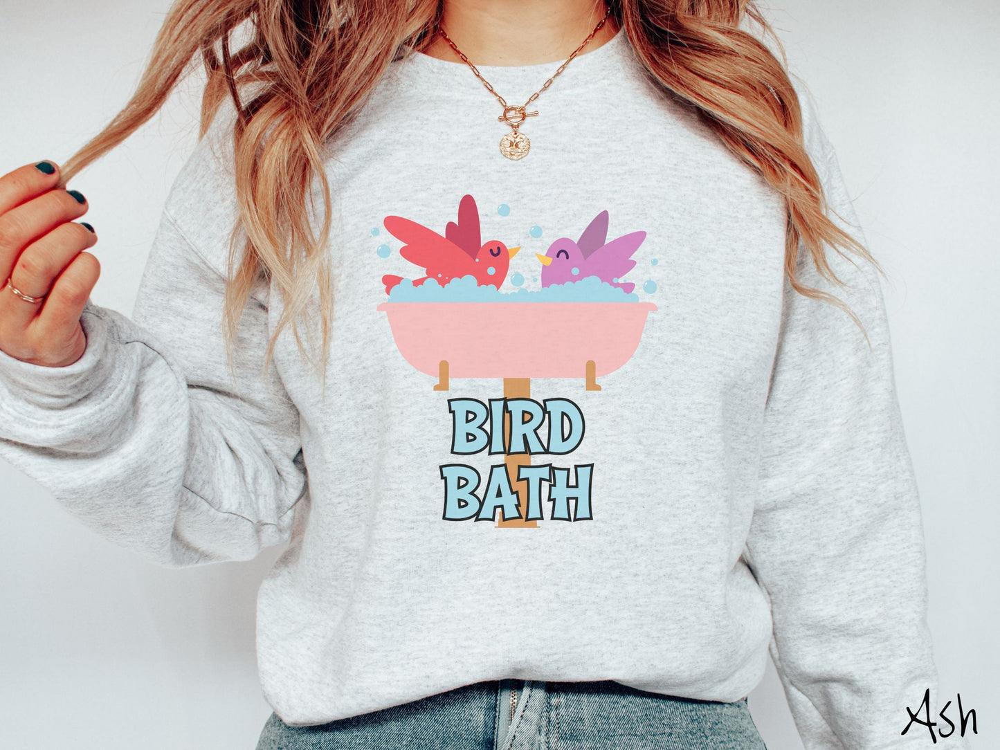 A woman wearing a cute, vintage ash colored comfy sweatshirt with a pink bathtub with a red bird and purple bird enjoying a bubble bath together. Below the bathtub are the words Bird Bath in light blue, uppercase font.