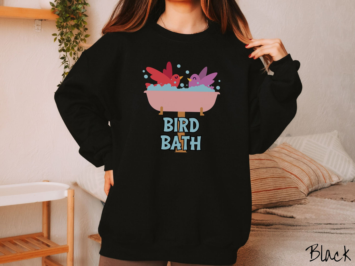 A woman wearing a cute, vintage black colored comfy sweatshirt with a pink bathtub with a red bird and purple bird enjoying a bubble bath together. Below the bathtub are the words Bird Bath in light blue, uppercase font.