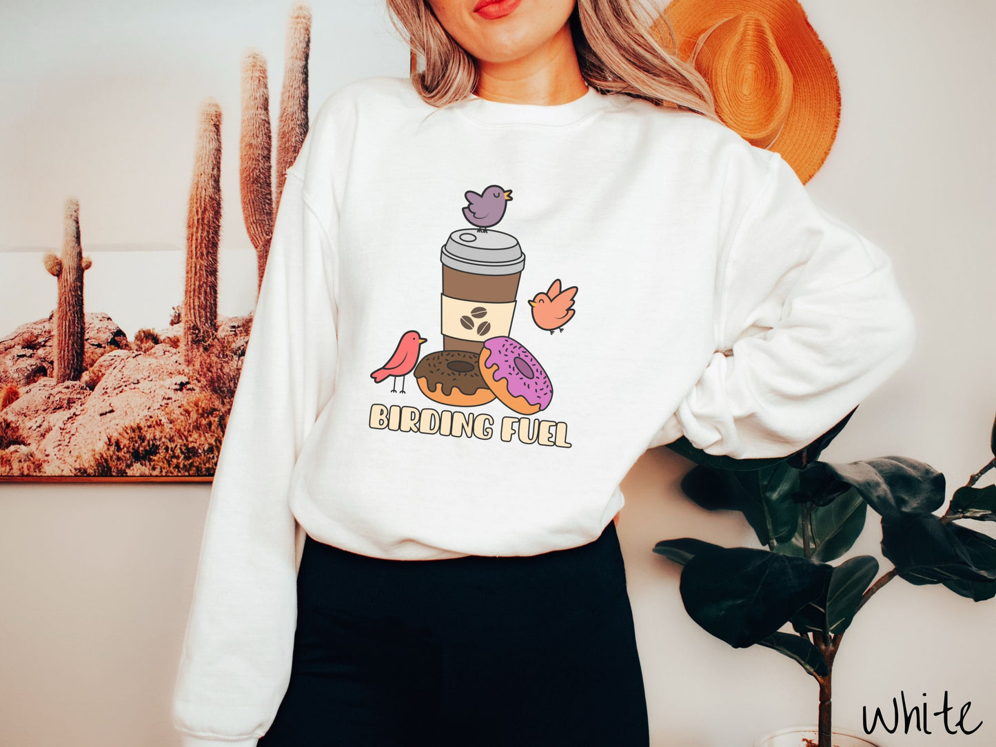 A woman wearing a cute, vintage white colored comfy sweatshirt with a brown to-go coffee cup next to a chocolate frosted donut and pink frosted donut. There are three orange, red, and purple birds sitting on the coffee cup and flying around.