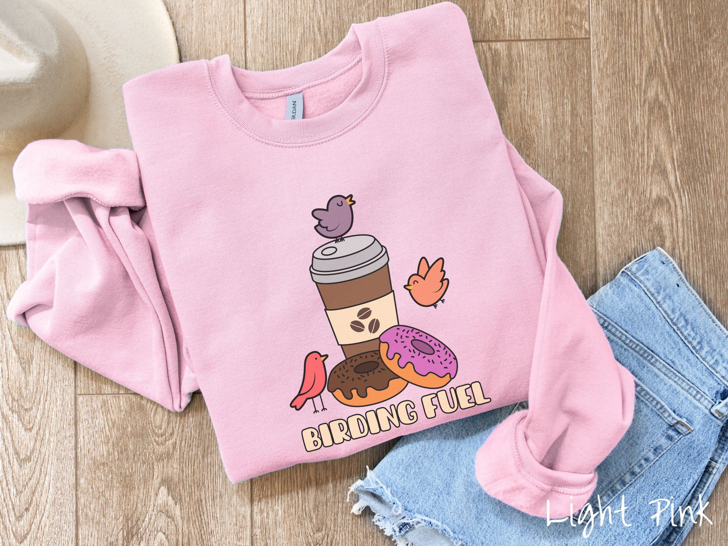 A cute, vintage light pinkcolored comfy sweatshirt with a brown to-go coffee cup next to a chocolate frosted donut and pink frosted donut. There are three orange, red, and purple birds sitting on the coffee cup and flying around.