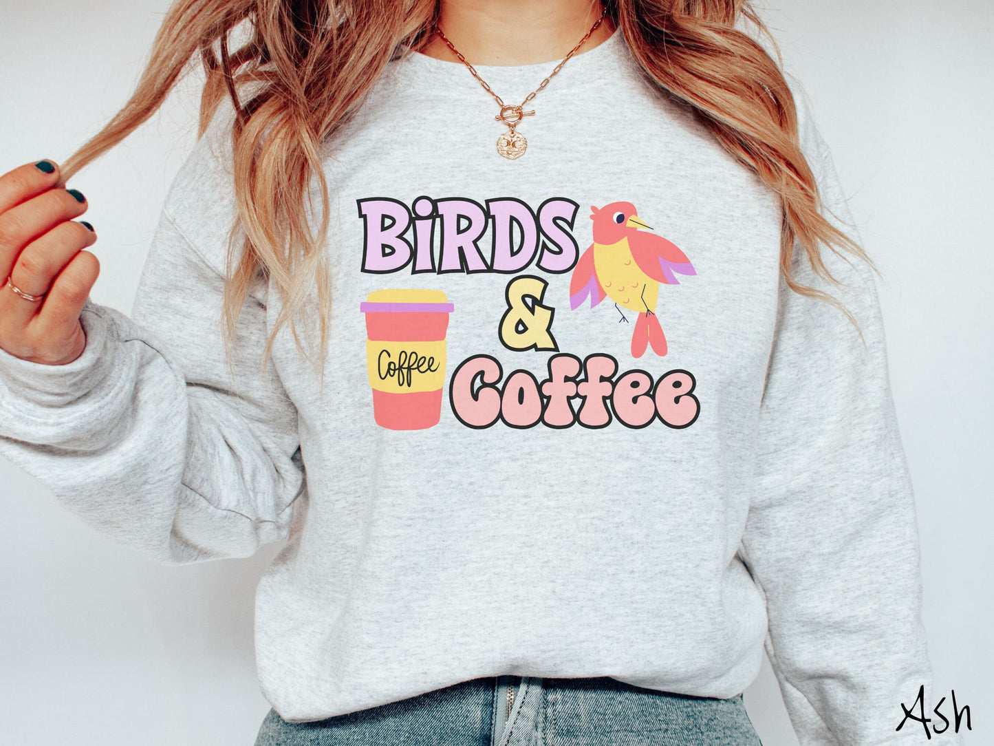 A woman wearing a cute, vintage ash colored comfy sweatshirt with the text Birds & Coffee in purple and light pink font. Next to the text is a red, yellow, and purple smiling bird in the air and a red and yellow to-go coffee cup.