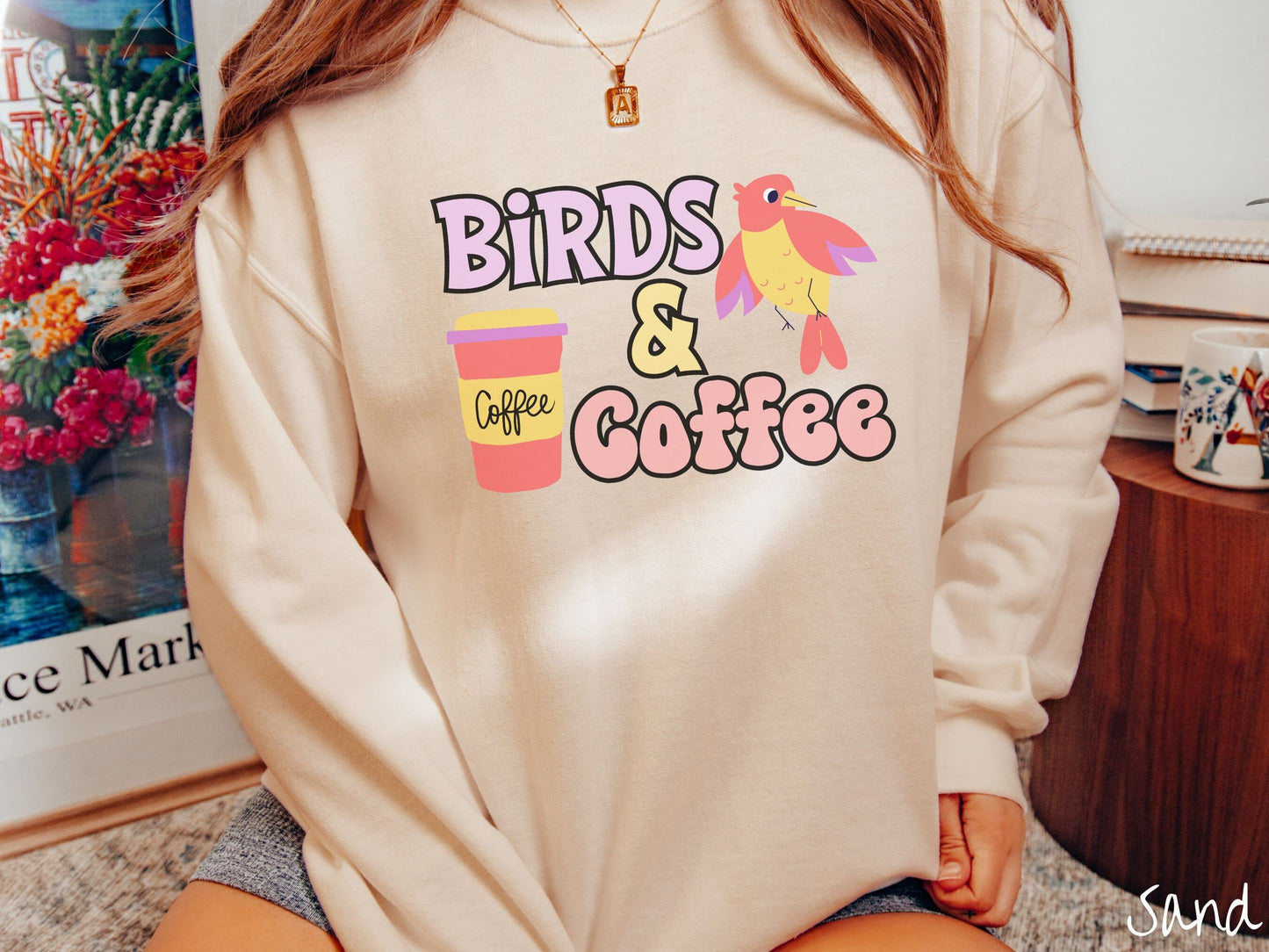 A woman wearing a cute, vintage sand colored comfy sweatshirt with the text Birds & Coffee in purple and light pink font. Next to the text is a red, yellow, and purple smiling bird in the air and a red and yellow to-go coffee cup.