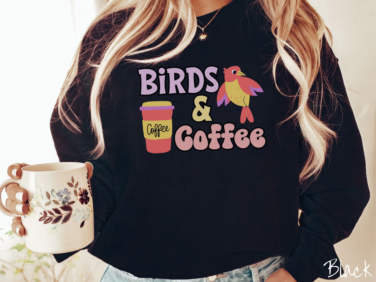 A woman wearing a cute, vintage black colored comfy sweatshirt with the text Birds & Coffee in purple and light pink font. Next to the text is a red, yellow, and purple smiling bird in the air and a red and yellow to-go coffee cup.