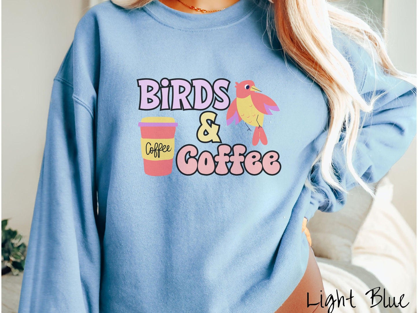 A woman wearing a cute, vintage light blue colored comfy sweatshirt with the text Birds & Coffee in purple and light pink font. Next to the text is a red, yellow, and purple smiling bird in the air and a red and yellow to-go coffee cup.