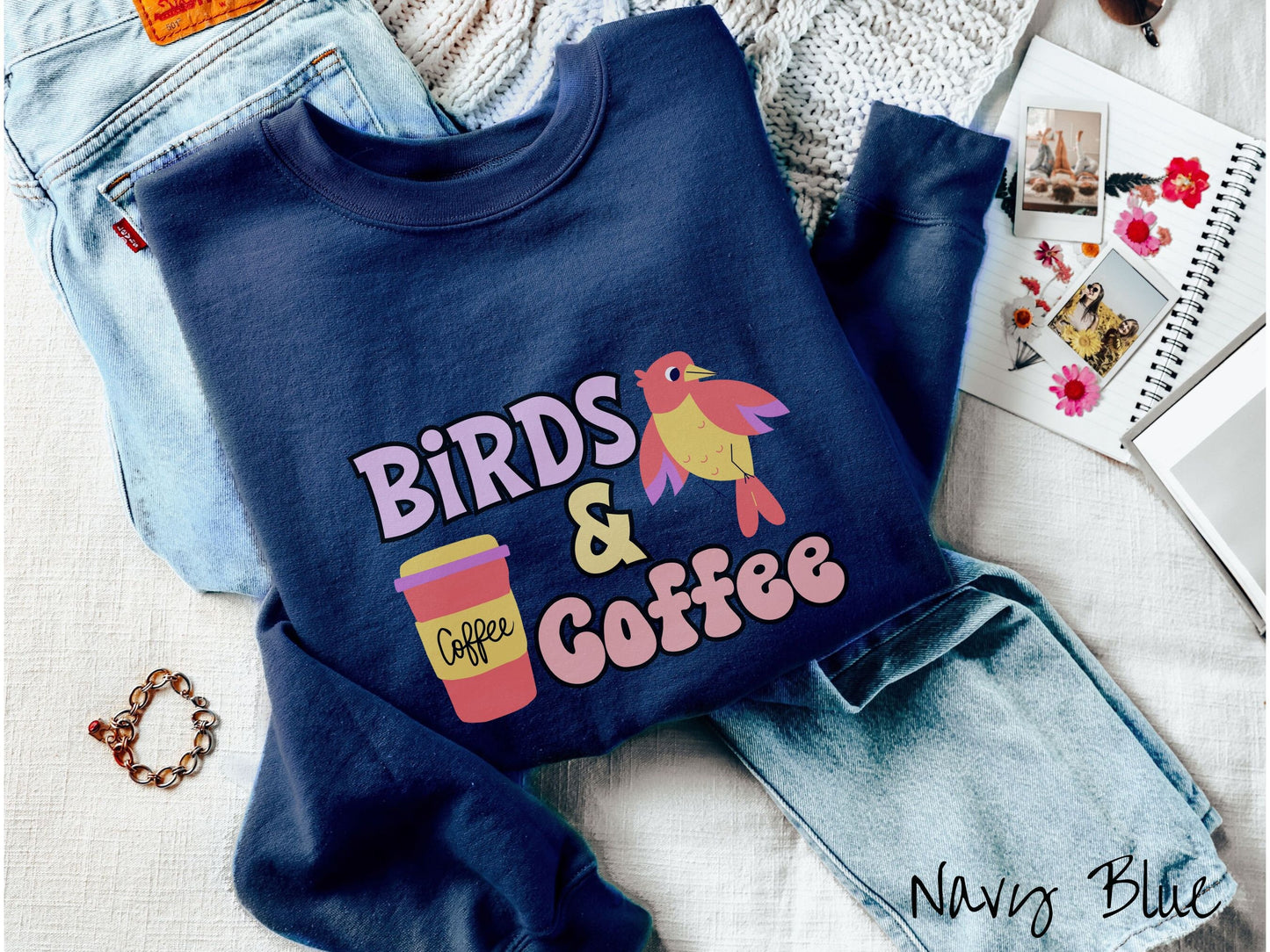 A cute, vintage navy blue colored comfy sweatshirt with the text Birds & Coffee in purple and light pink font. Next to the text is a red, yellow, and purple smiling bird in the air and a red and yellow to-go coffee cup.