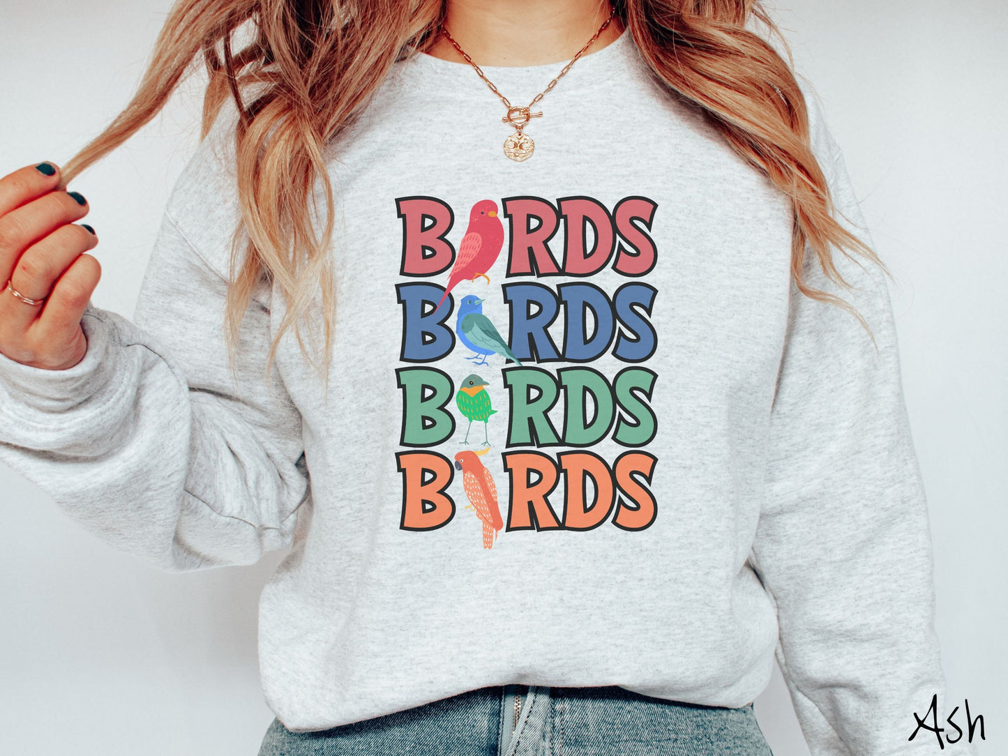 A woman wearing a cute, vintage ash colored comfy sweatshirt with the word BIRDS listed four times vertically in red, blue, green, and orange uppercase font. The Is in BIRDS are different kinds of colorful birds.