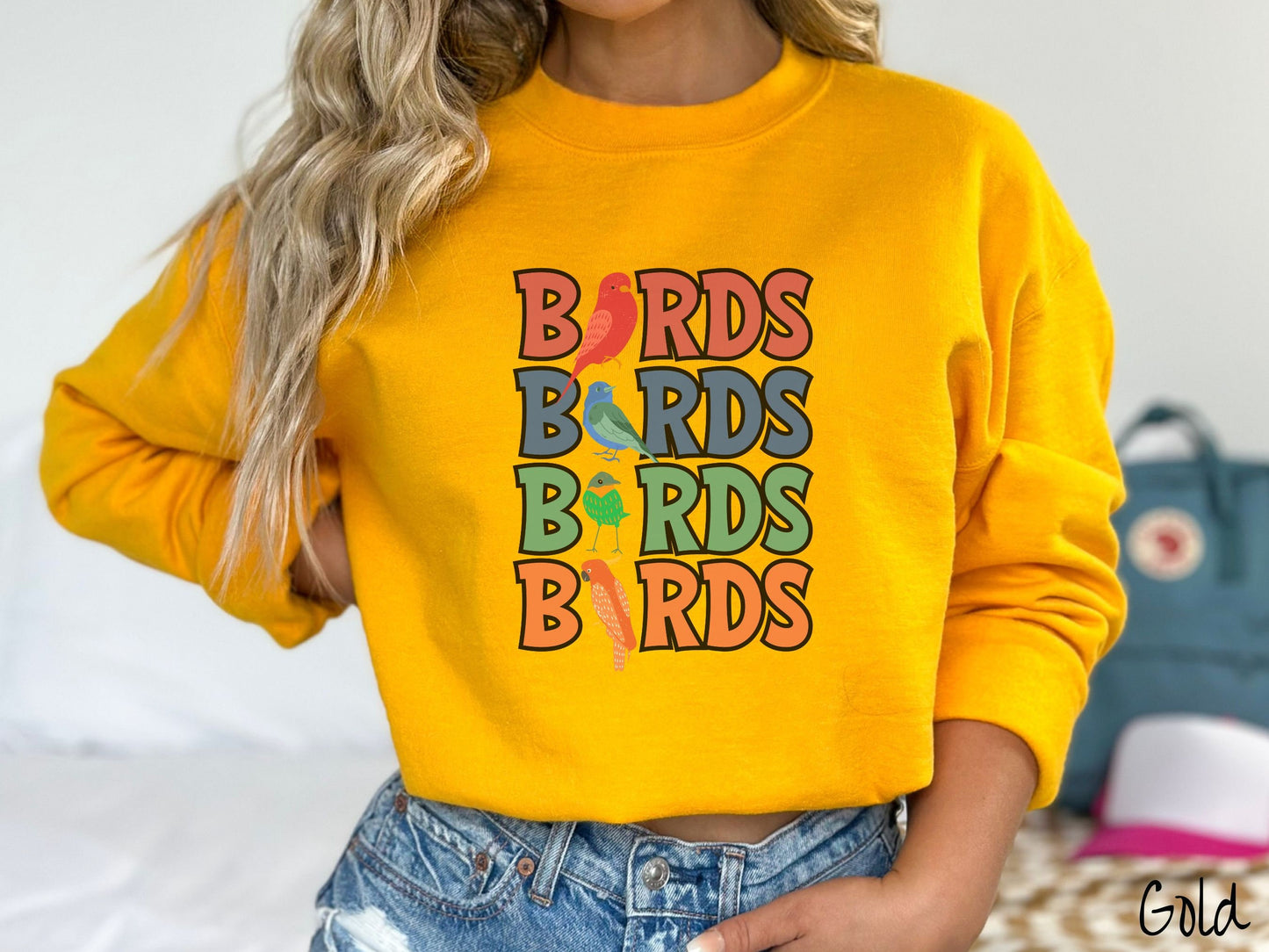 A woman wearing a cute, vintage gold colored comfy sweatshirt with the word BIRDS listed four times vertically in red, blue, green, and orange uppercase font. The Is in BIRDS are different kinds of colorful birds.