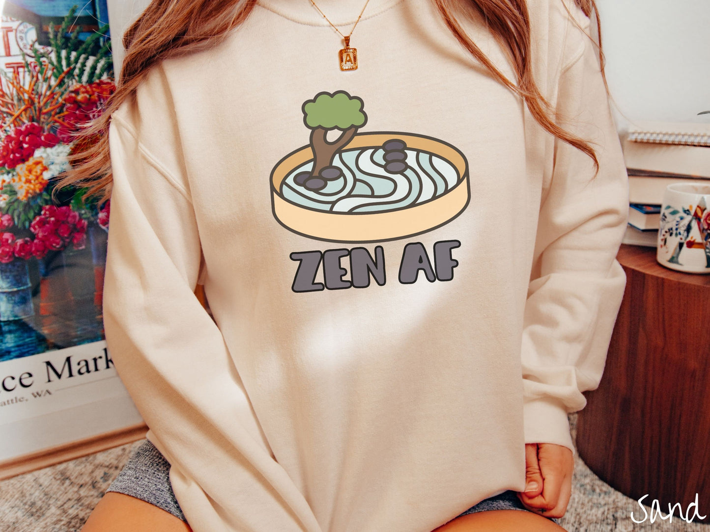 A woman wearing a cute, vintage sand colored comfy sweatshirt with the text Zen AF in gray font across the font. Above this is a zen garden in a yellow container with gray and light green sand, some black pebbles, and a green tree.