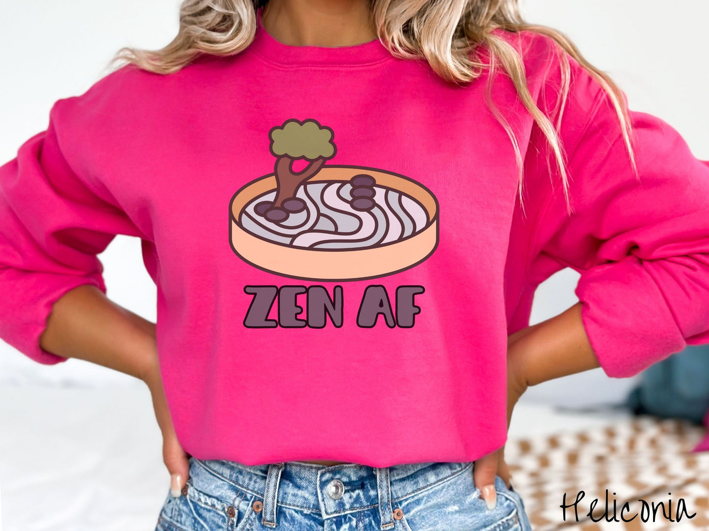A woman wearing a cute, vintage heliconia colored comfy sweatshirt with the text Zen AF in gray font across the font. Above this is a zen garden in a yellow container with gray and light green sand, some black pebbles, and a green tree.