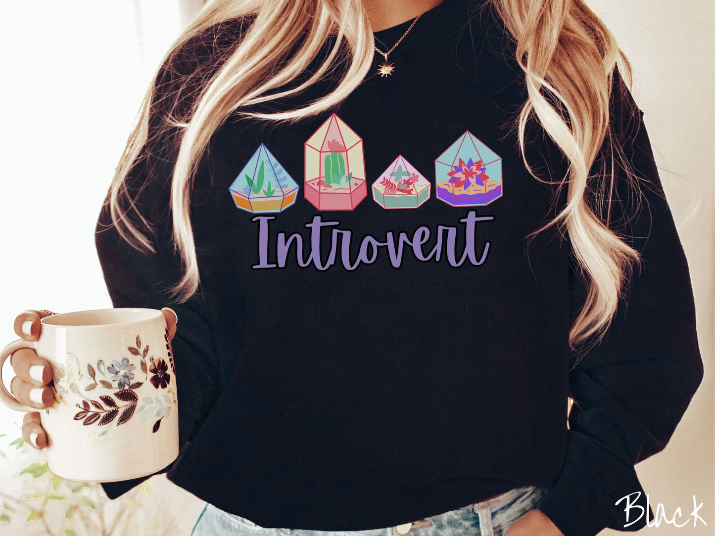 A woman wearing a cute, vintage black colored comfy sweatshirt with the text Introvert in purple font across the front. Above the text are four colorful plant terrariums with colorful flowers and cacti inside.