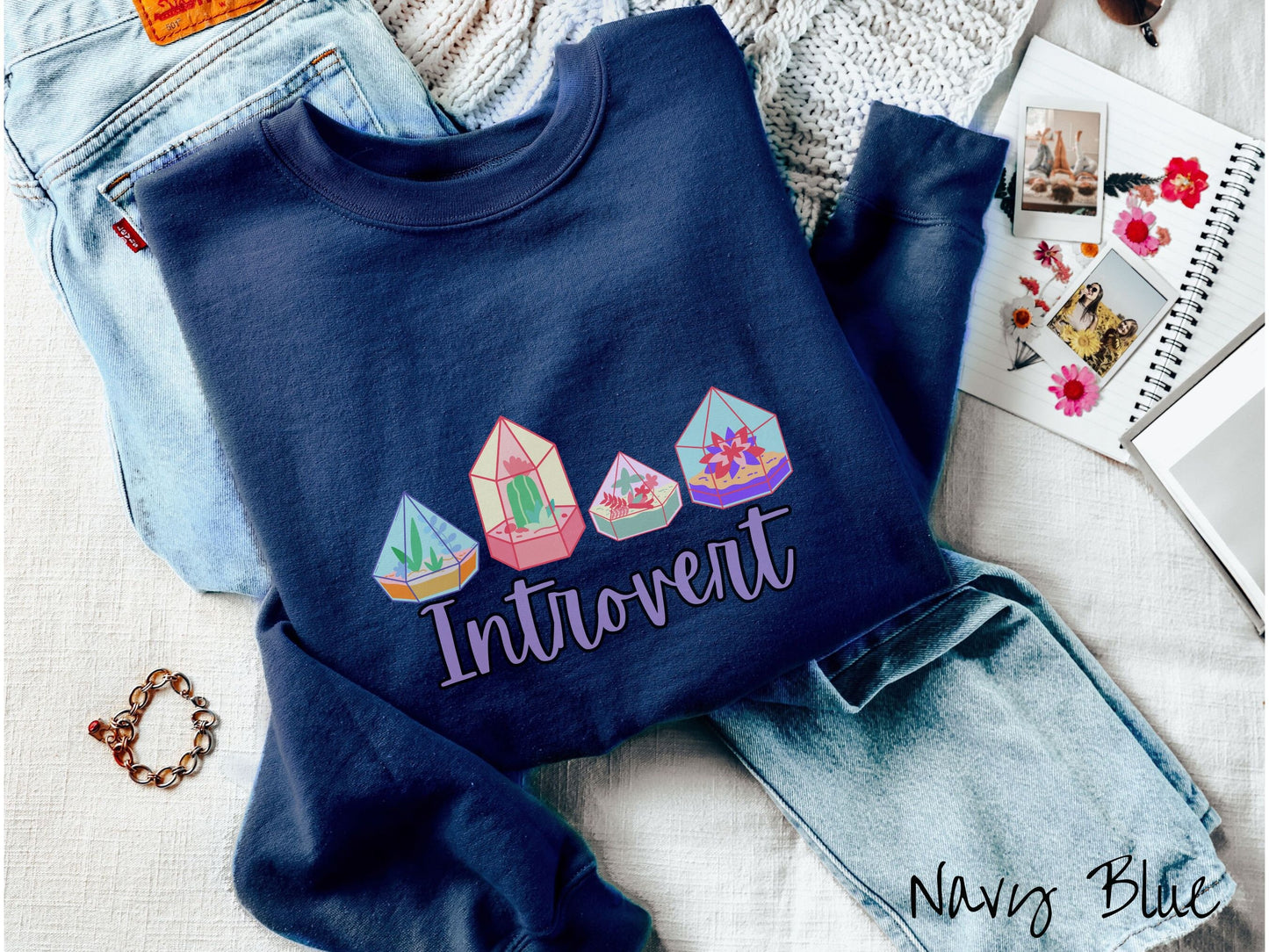 A cute, vintage navy blue colored comfy sweatshirt with the text Introvert in purple font across the front. Above the text are four colorful plant terrariums with colorful flowers and cacti inside.