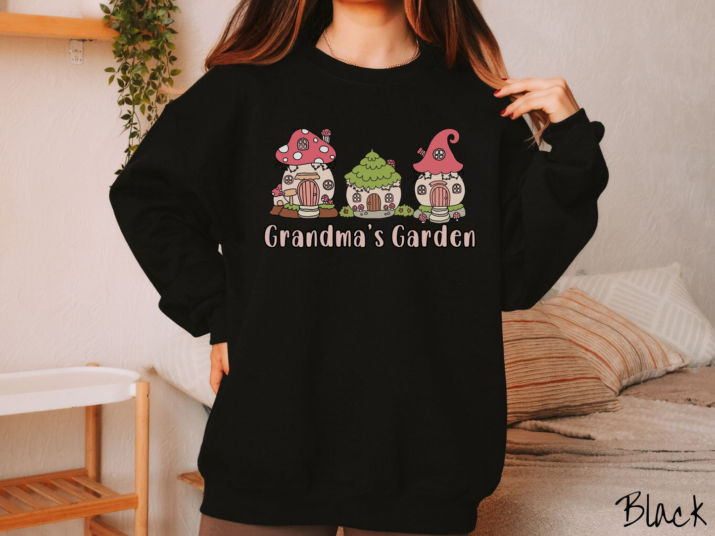 A woman wearing a cute, vintage black colored comfy sweatshirt with the text Grandmas Garden in light pink font. Above the text are cute little cottages for gnomes made from light and dark pink mushrooms.
