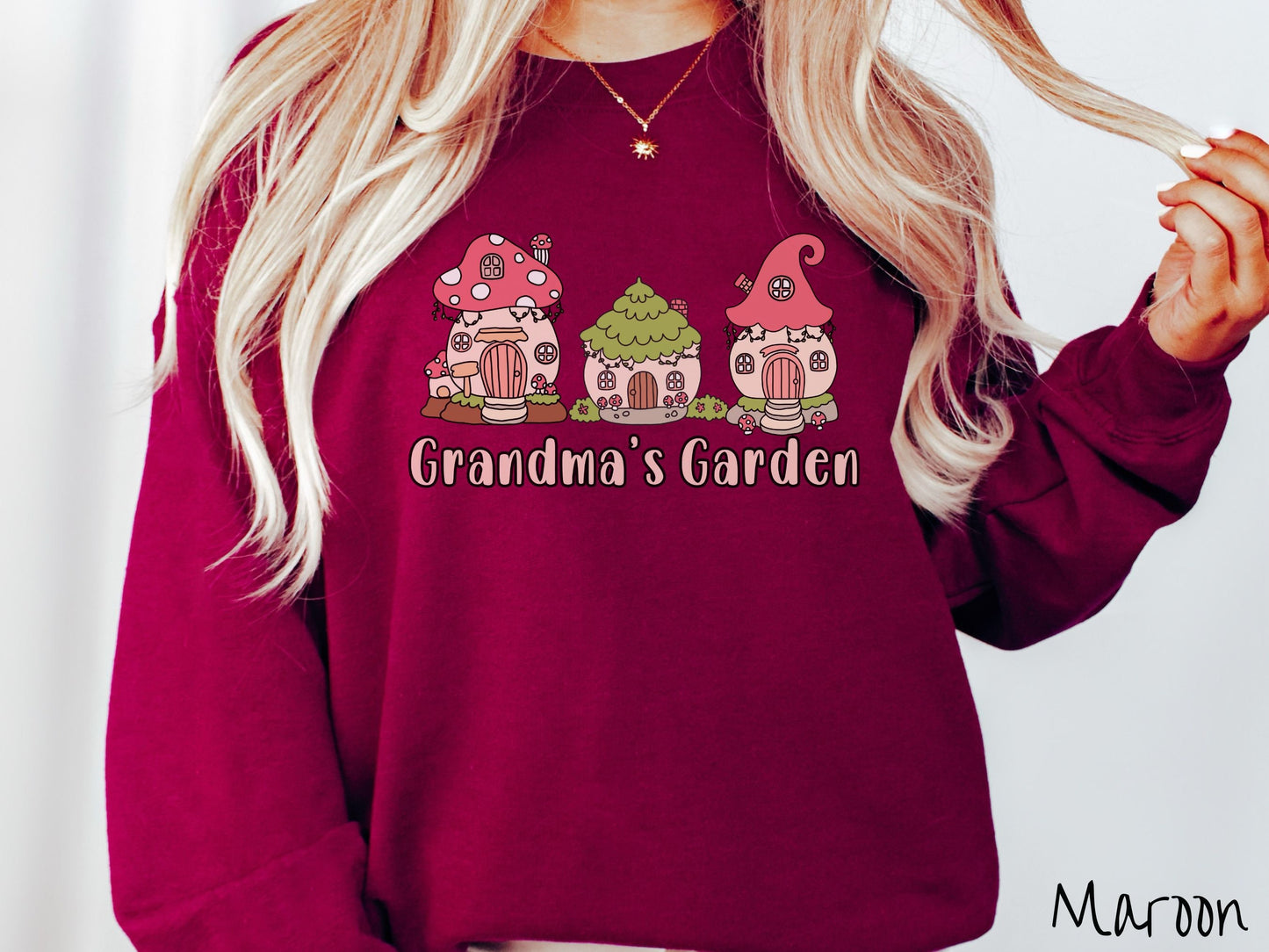 A woman wearing a cute, vintage maroon colored comfy sweatshirt with the text Grandmas Garden in light pink font. Above the text are cute little cottages for gnomes made from light and dark pink mushrooms.