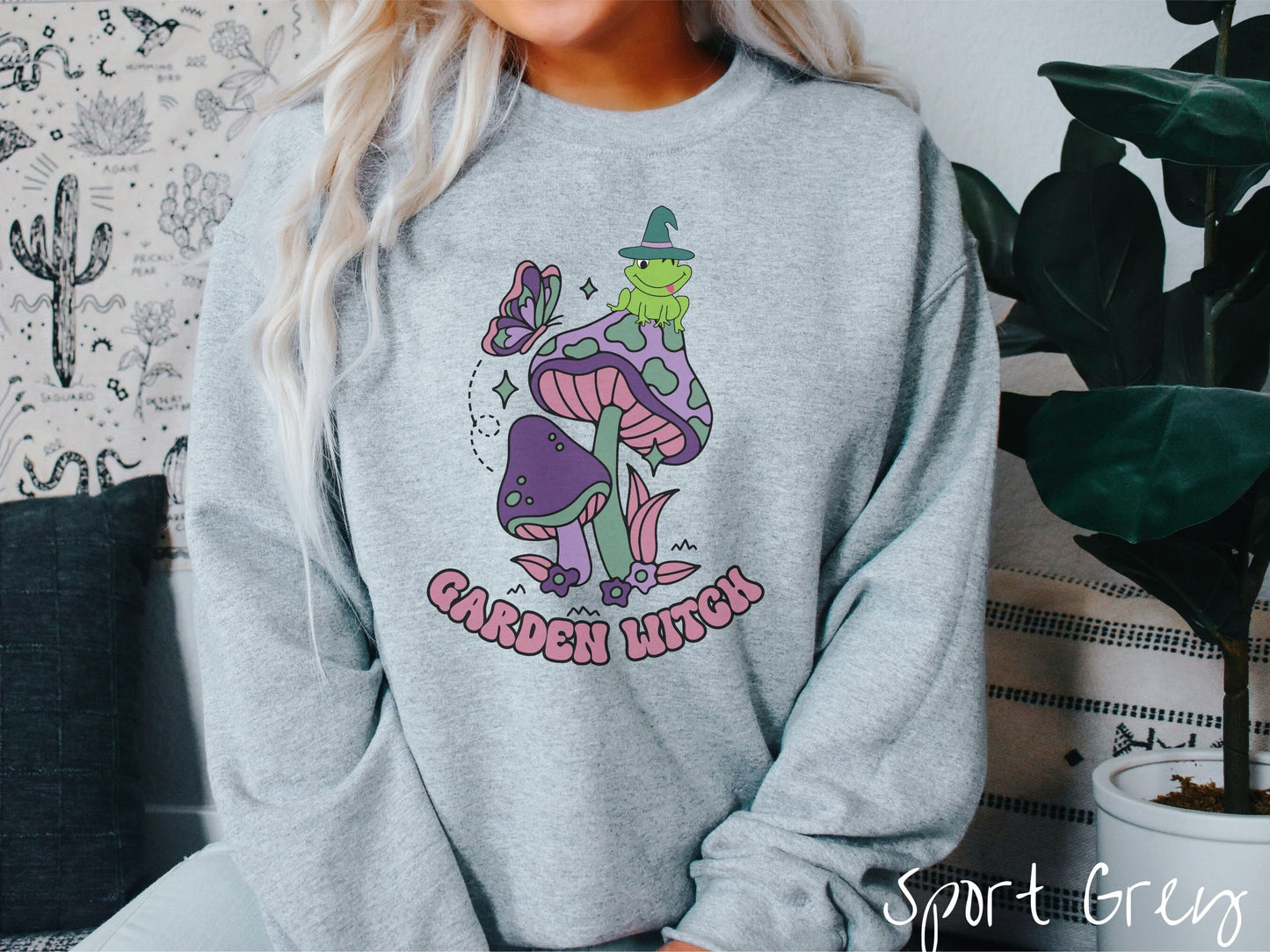 A woman wearing a cute, vintage sport grey colored comfy sweatshirt with the text Garden Witch in purple font. Above this text are two purple and green mushrooms with a green frog sitting atop one wearing a witch hat.
