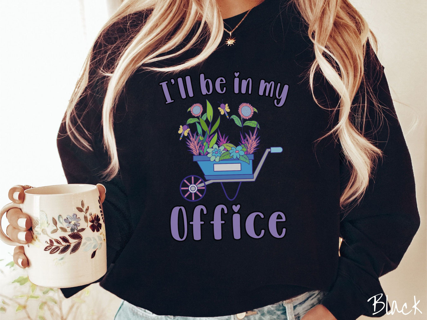 A woman wearing a cute, vintage black colored comfy sweatshirt with the text I’ll Be in My Office in purple font across the front. In between the text is a blue wheelbarrow with a pink wheel stacked full of colorful plants and flowers.