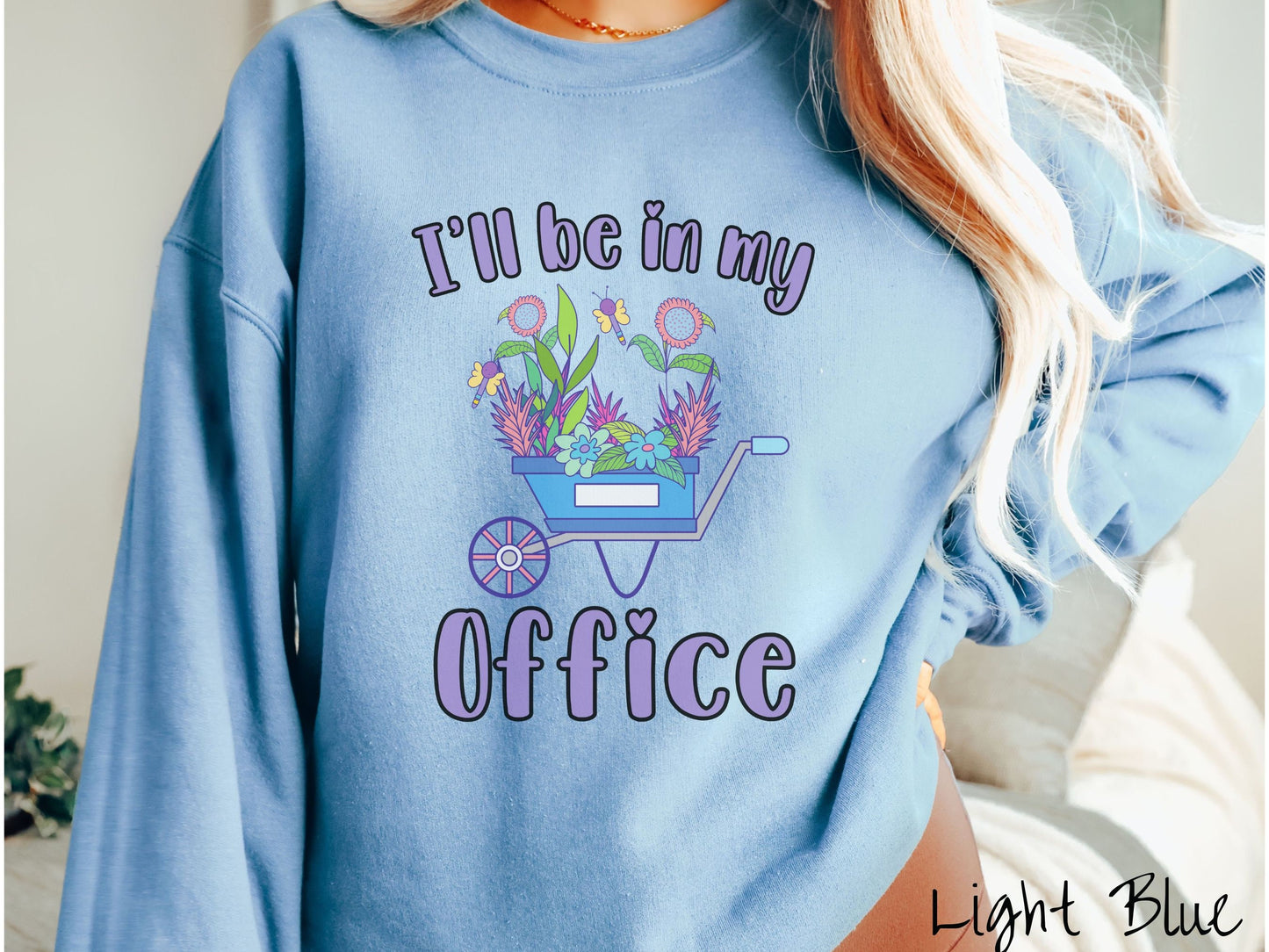 A woman wearing a cute, vintage light blue colored comfy sweatshirt with the text I’ll Be in My Office in purple font across the front. In between the text is a blue wheelbarrow with a pink wheel stacked full of colorful plants and flowers.