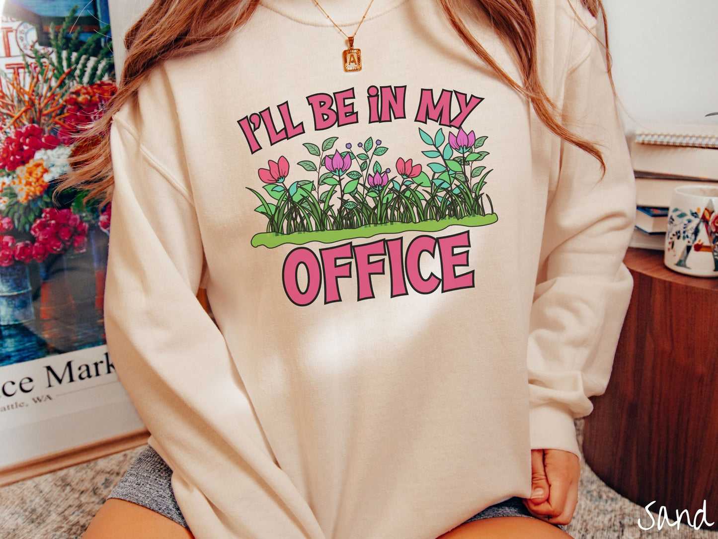 A woman wearing a cute, vintage sand colored comfy sweatshirt with the text I’ll Be in my Office in red font across the front. In between the text is a garden of red and pink flowers, green plants, and lush green grass.
