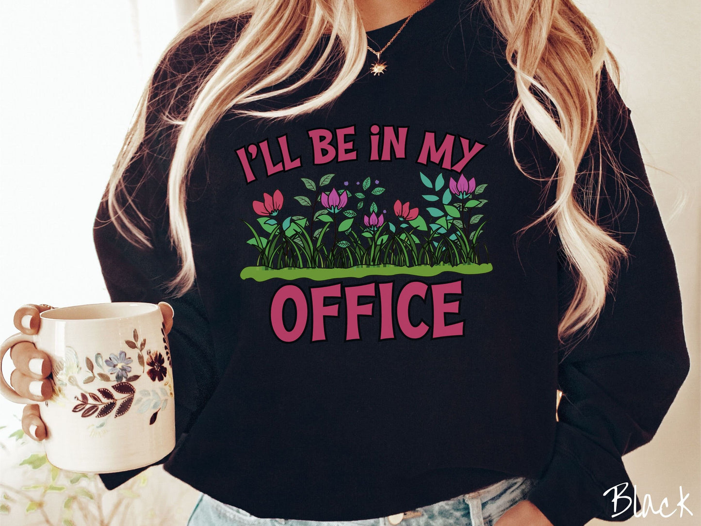 A woman wearing a cute, vintage black colored comfy sweatshirt with the text I’ll Be in my Office in red font across the front. In between the text is a garden of red and pink flowers, green plants, and lush green grass.
