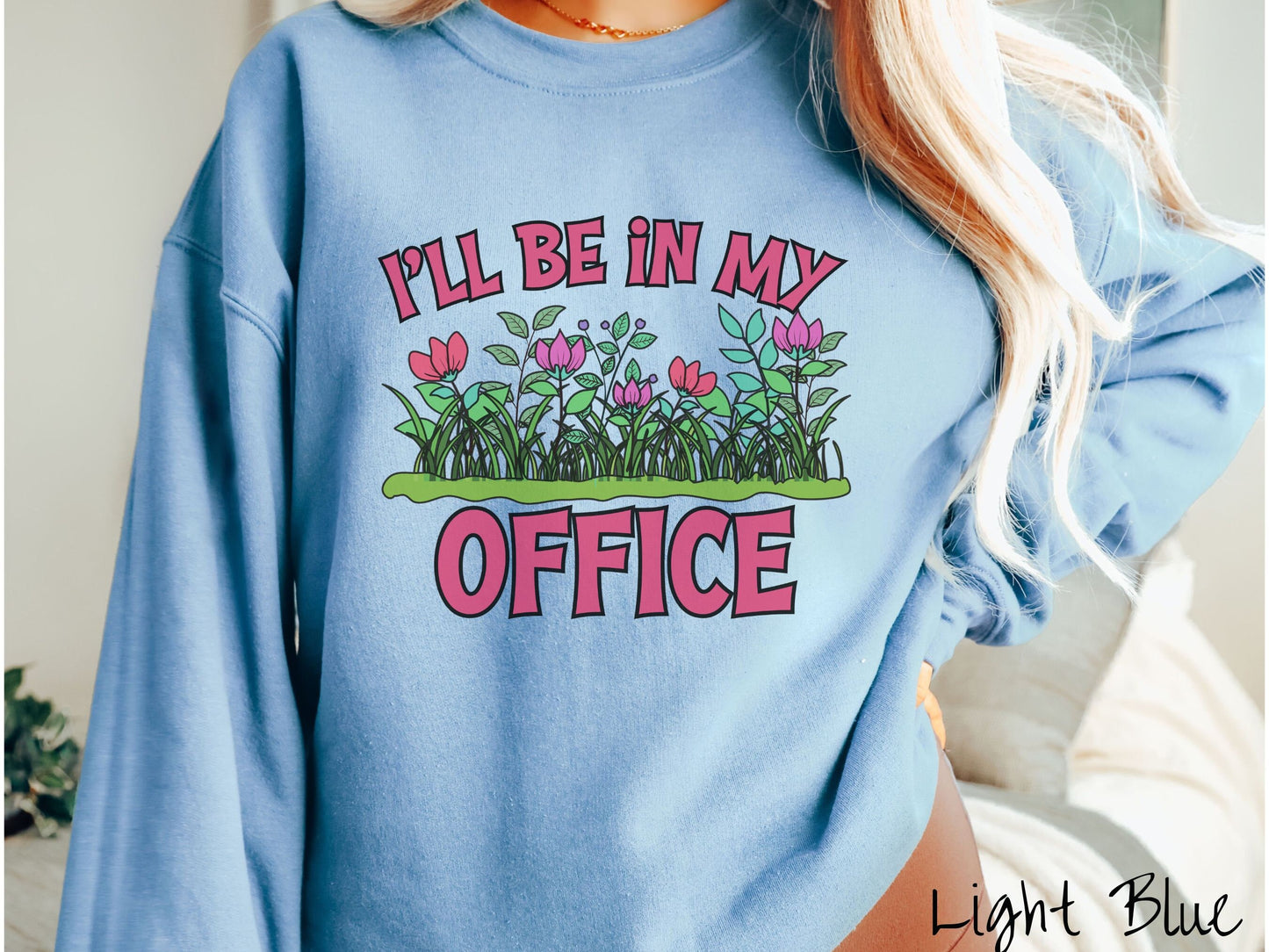 A woman wearing a cute, vintage light blue colored comfy sweatshirt with the text I’ll Be in my Office in red font across the front. In between the text is a garden of red and pink flowers, green plants, and lush green grass.