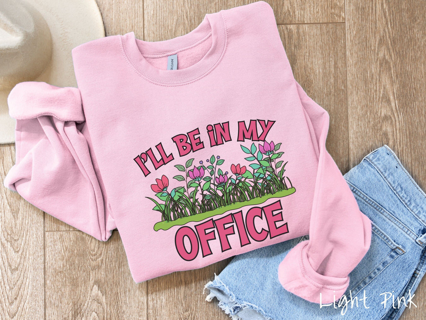 A cute, vintage light pink colored comfy sweatshirt with the text I’ll Be in my Office in red font across the front. In between the text is a garden of red and pink flowers, green plants, and lush green grass.