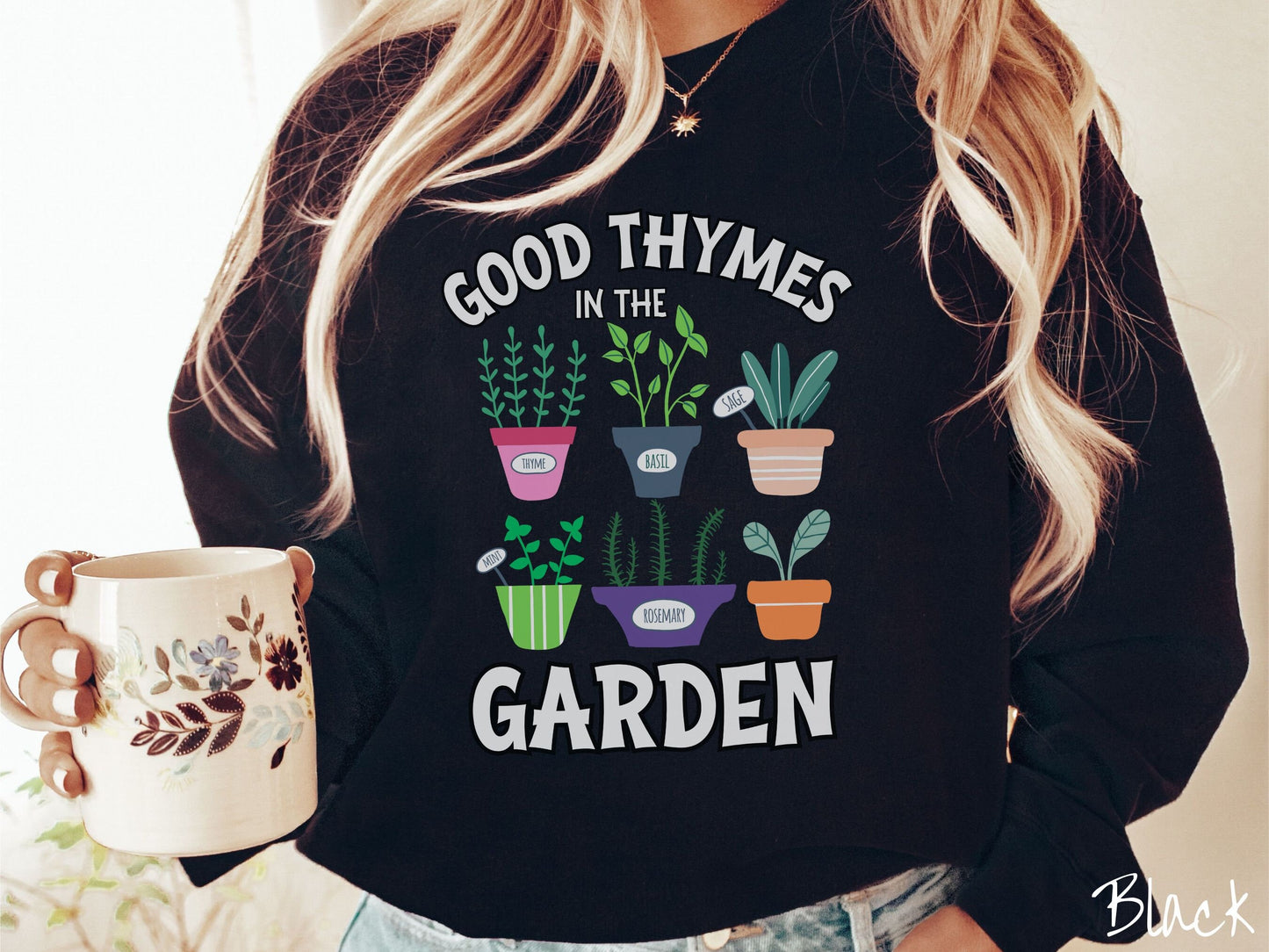 A woman wearing a cute, vintage black colored comfy sweatshirt with the text Good Thymes in the Garden in white font. In between the text are different potted vegetables in colorful pots like thyme, basil, sage, and mint.