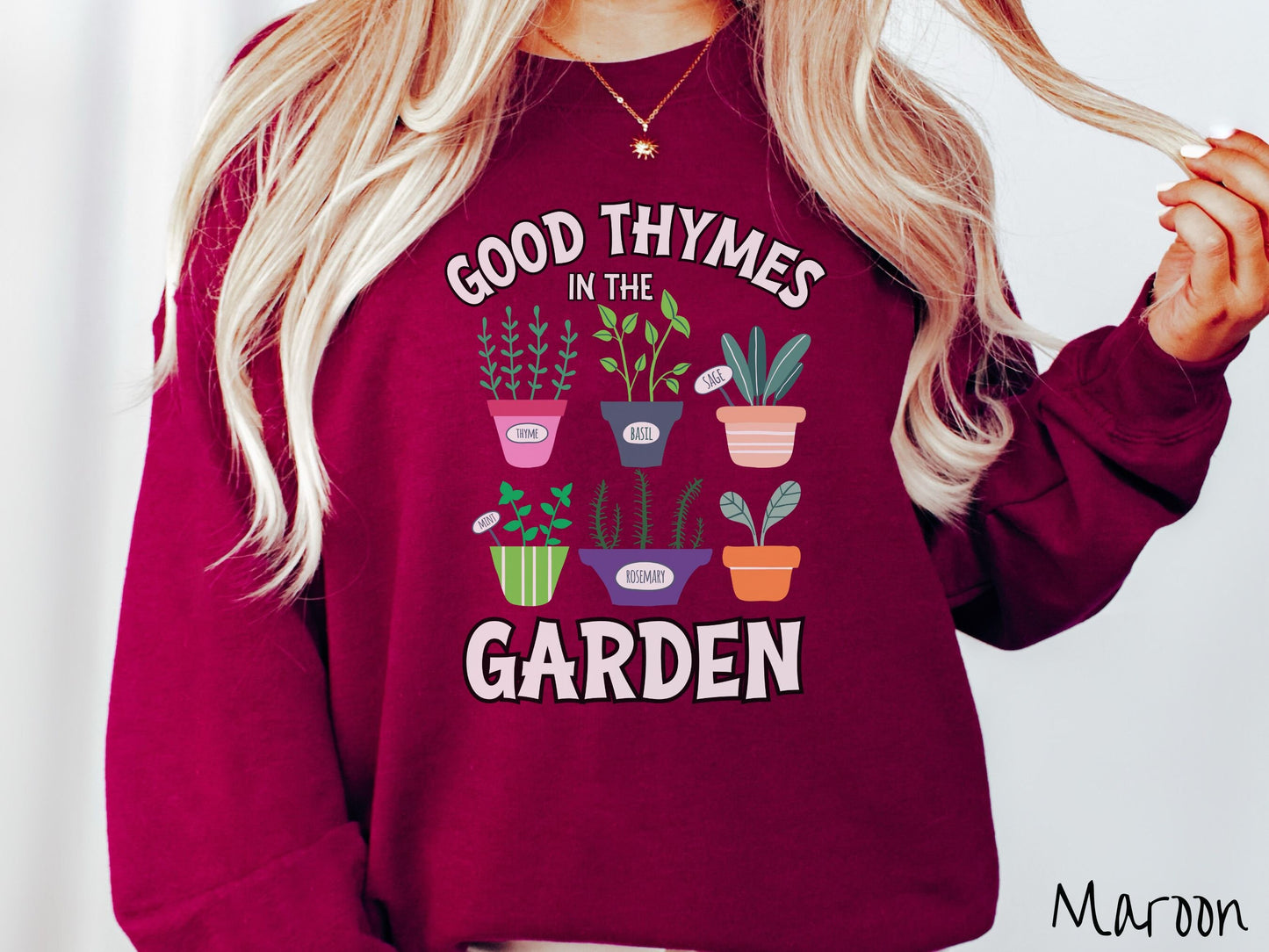 A woman wearing a cute, vintage maroon colored comfy sweatshirt with the text Good Thymes in the Garden in white font. In between the text are different potted vegetables in colorful pots like thyme, basil, sage, and mint.