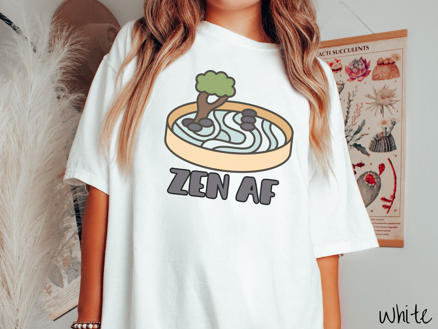A woman wearing a cute, vintage white colored Comfort Colors t-shirt with the text Zen AF in gray font across the font. Above this is a zen garden in a yellow container with gray and light green sand, some black pebbles, and a green tree.