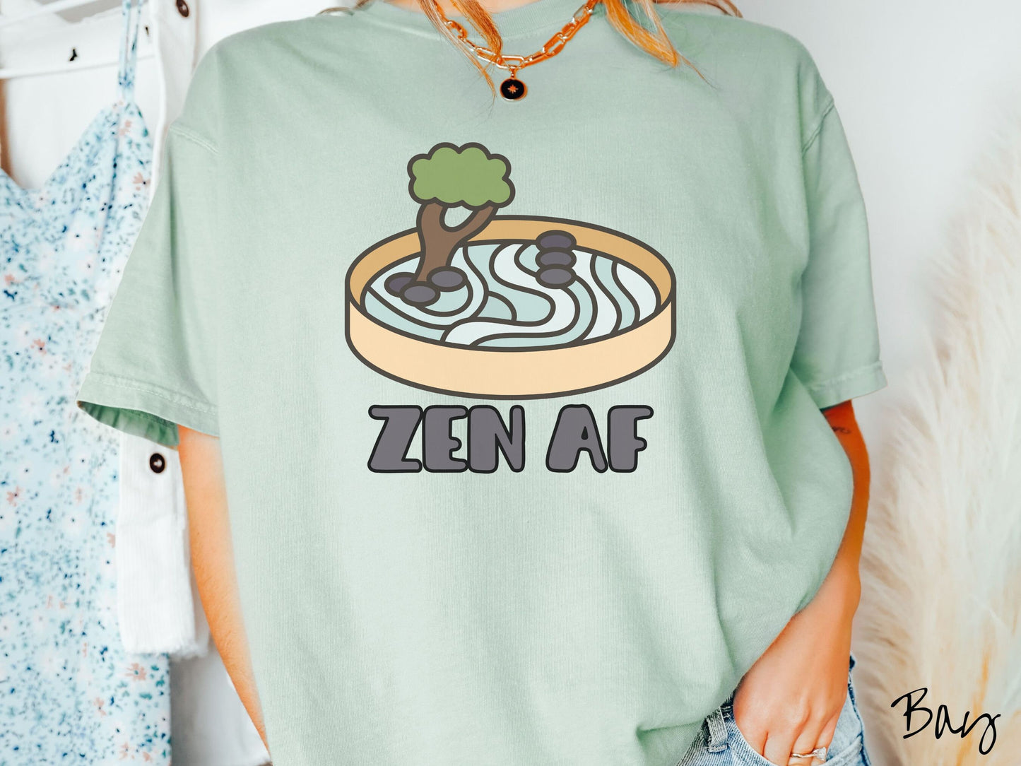 A woman wearing a cute, vintage bay colored Comfort Colors t-shirt with the text Zen AF in gray font across the font. Above this is a zen garden in a yellow container with gray and light green sand, some black pebbles, and a green tree.