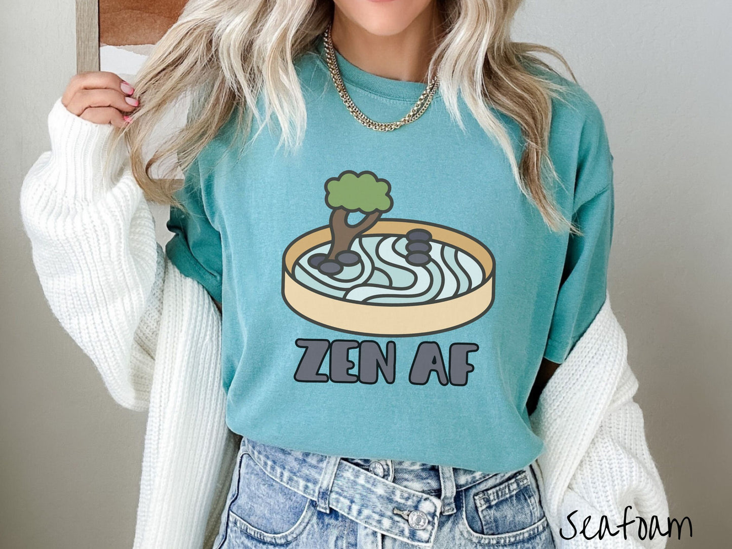 A woman wearing a cute, vintage seafoam colored Comfort Colors t-shirt with the text Zen AF in gray font across the font. Above this is a zen garden in a yellow container with gray and light green sand, some black pebbles, and a green tree.