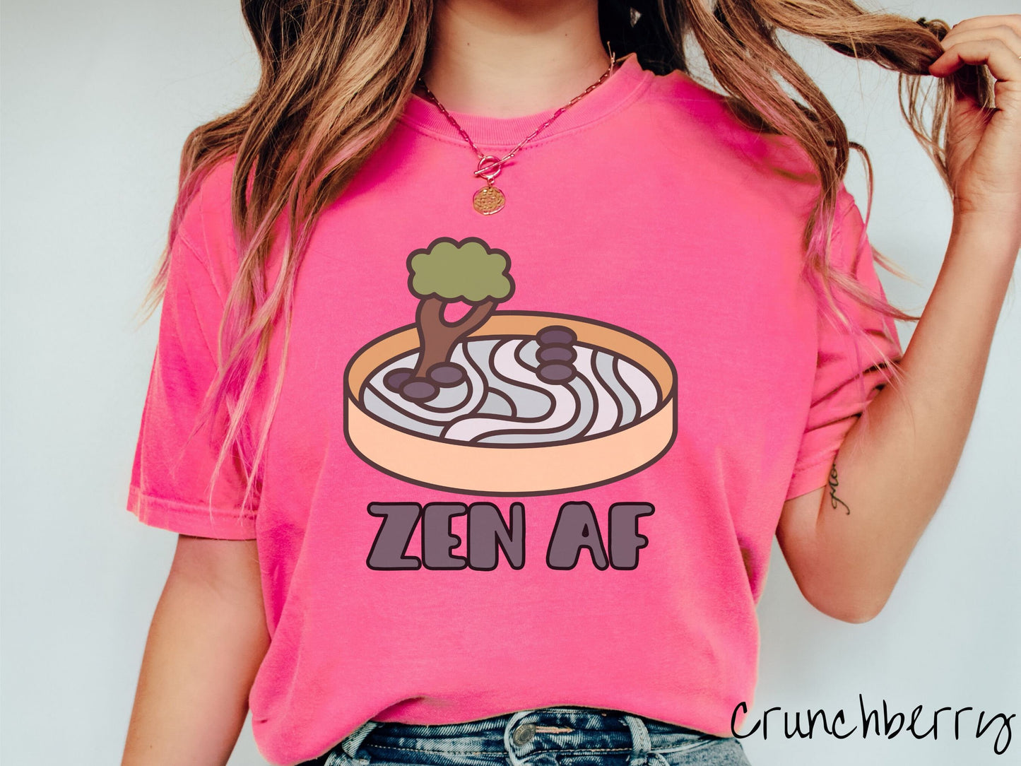 A woman wearing a cute, vintage crunchberry colored Comfort Colors t-shirt with the text Zen AF in gray font across the font. Above this is a zen garden in a yellow container with gray and light green sand, some black pebbles, and a green tree.